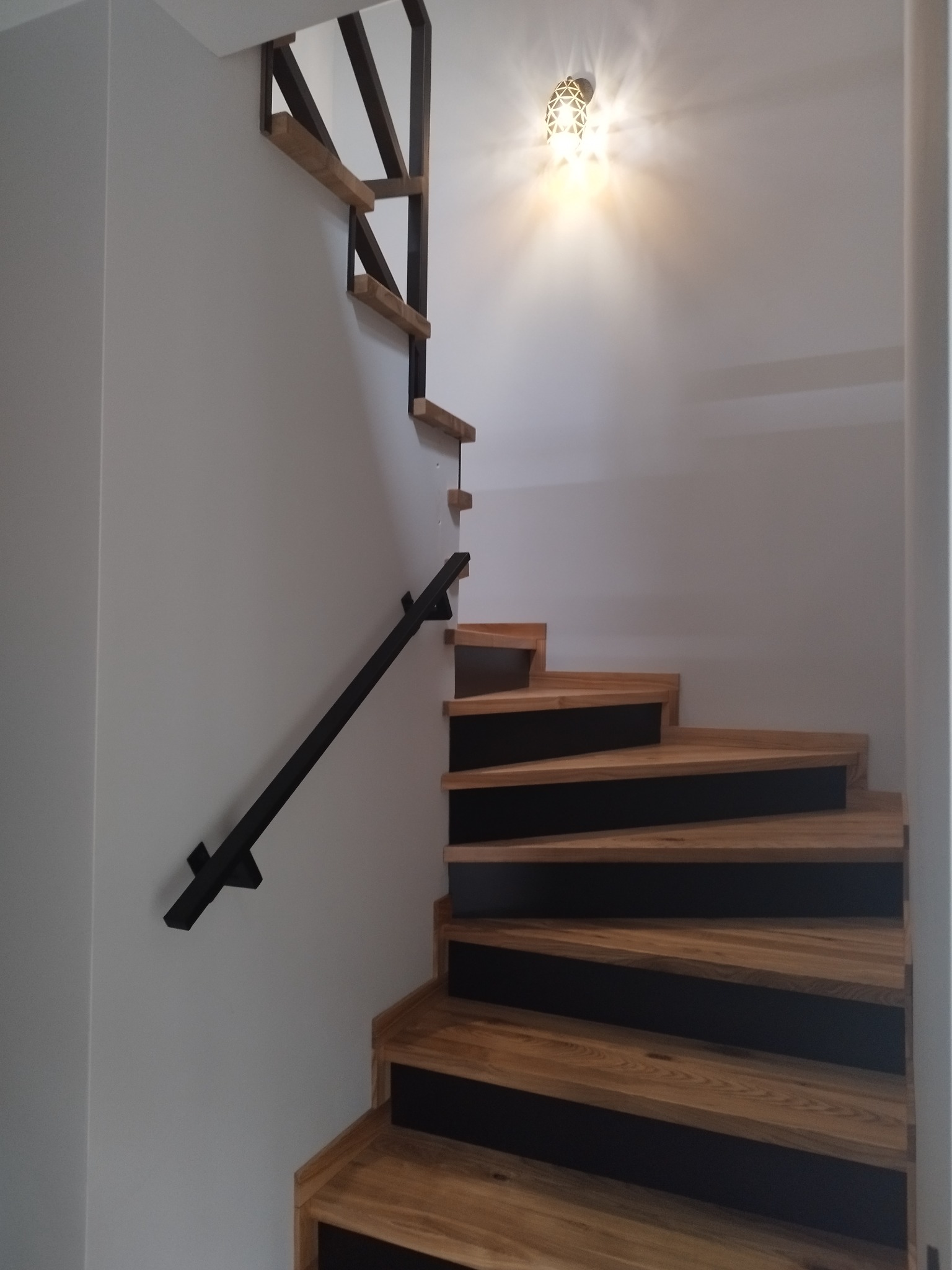 Nowoczesne schody drewniane z czarnymi podstopnicami i metalową balustradą, oświetlone dekoracyjną lampą na suficie.
