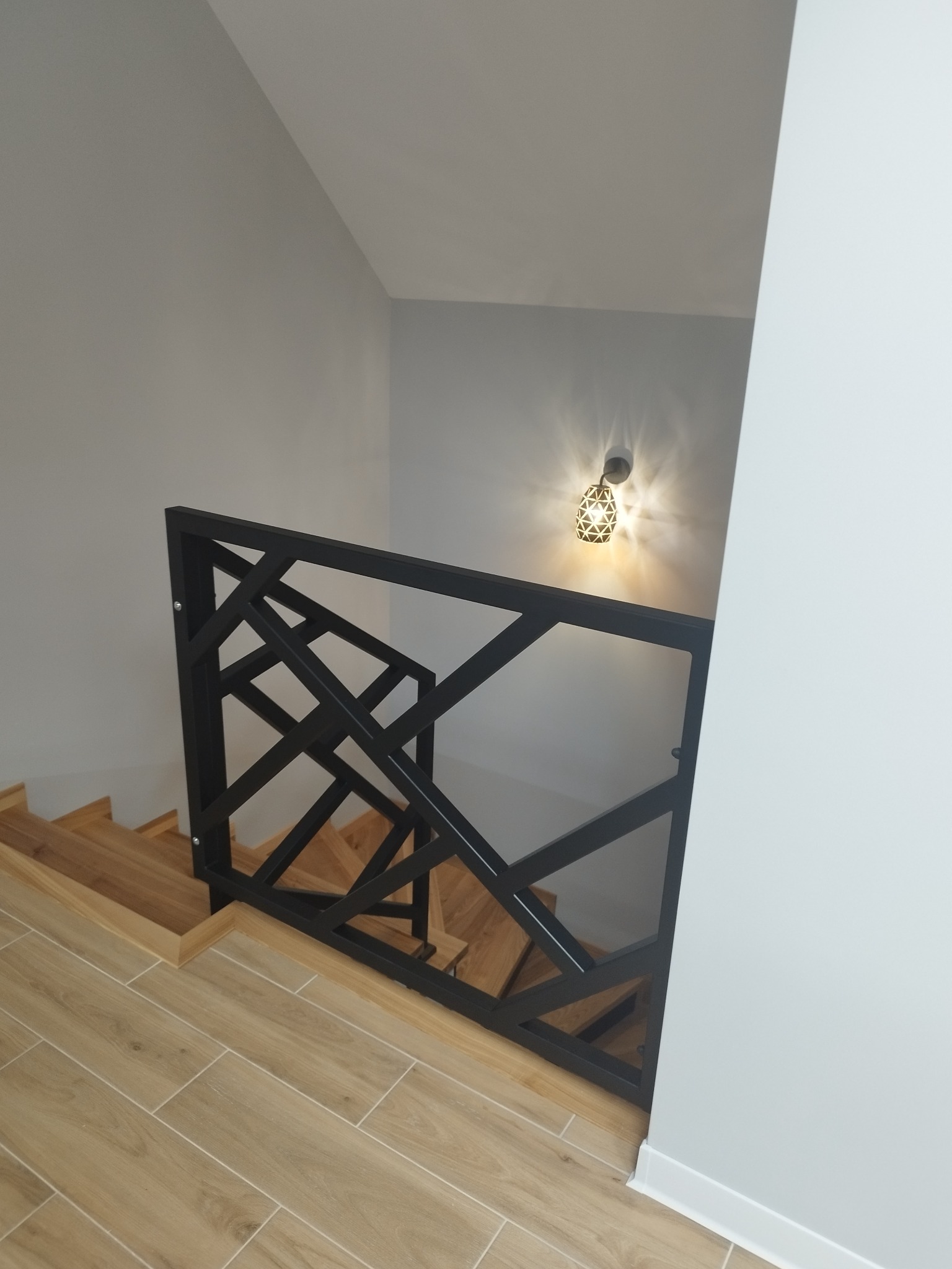 Nowoczesna balustrada schodowa z czarnego metalu o geometrycznym wzorze w Łomży, zamontowana na drewnianych schodach w jasnym wnętrzu.