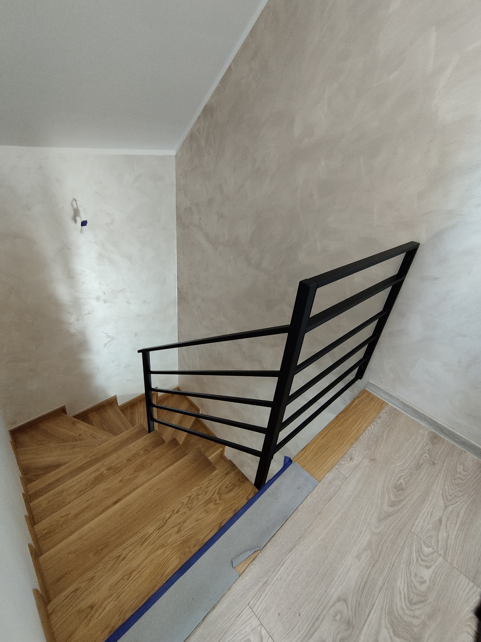 Kątowe ujęcie drewnianych schodów z czarną, minimalistyczną balustradą, ściany wykończone w odcieniach beżu, widok z góry.