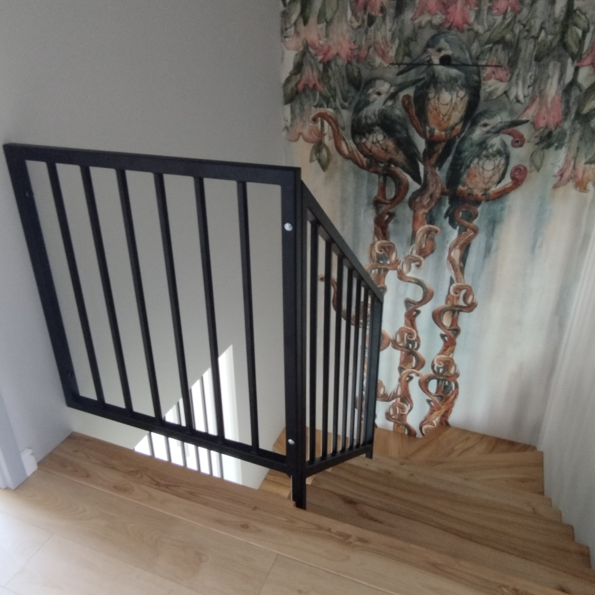 Fragment czarnej, metalowej balustrady schodowej z pionowymi szczebelkami zamocowanej do drewnianych schodów, na ścianie widoczna tapeta z motywem ptaków i kwiatów.