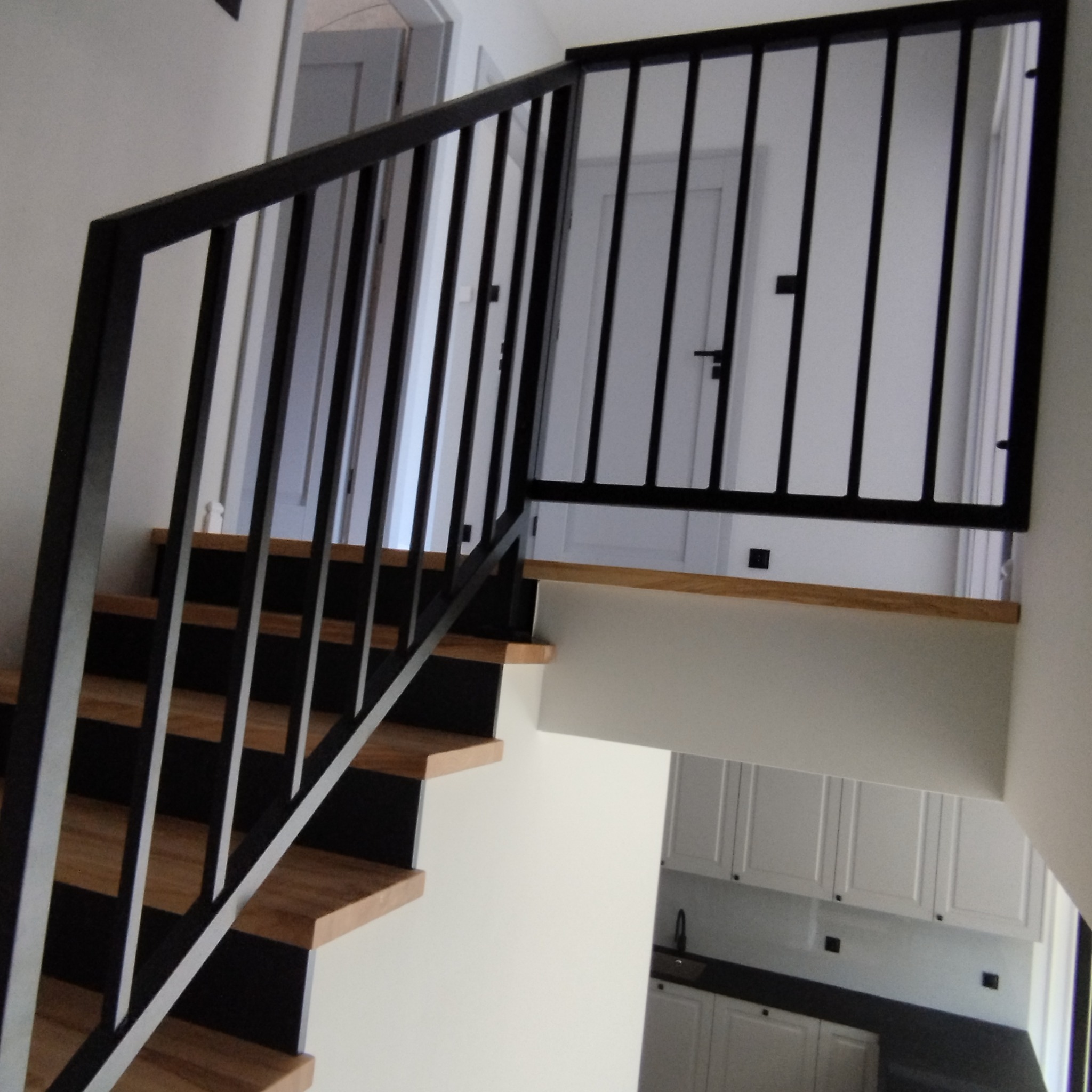 Nowoczesne schody z drewnianymi stopniami i czarną, metalową balustradą o pionowych szczeblach, widziane z dołu, z fragmentem białej kuchni w tle.