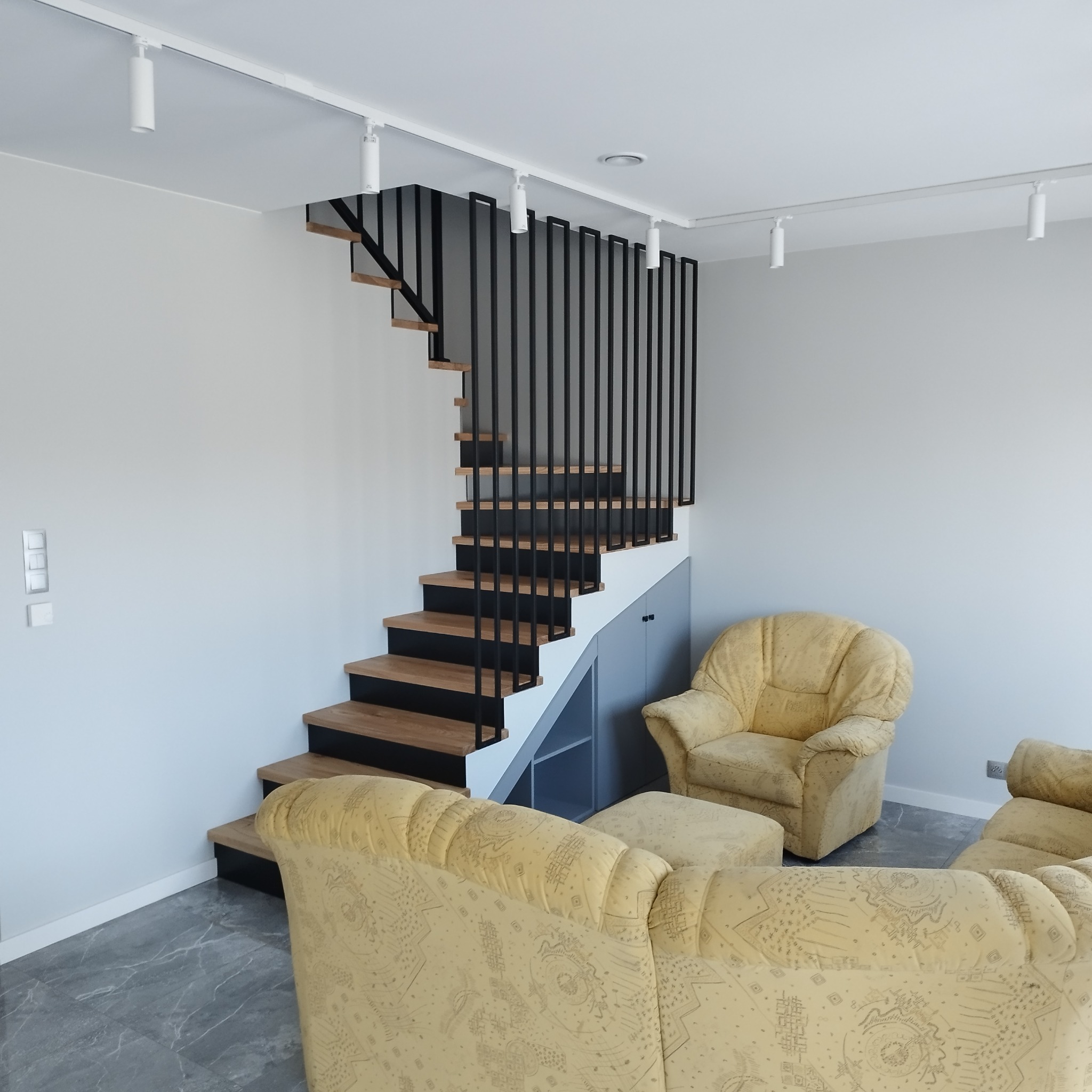 Nowoczesne schody metalowe z drewnianymi stopniami w jasnym wnętrzu, z czarną balustradą pionową i oświetleniem punktowym na suficie, widoczne meble wypoczynkowe w tle.