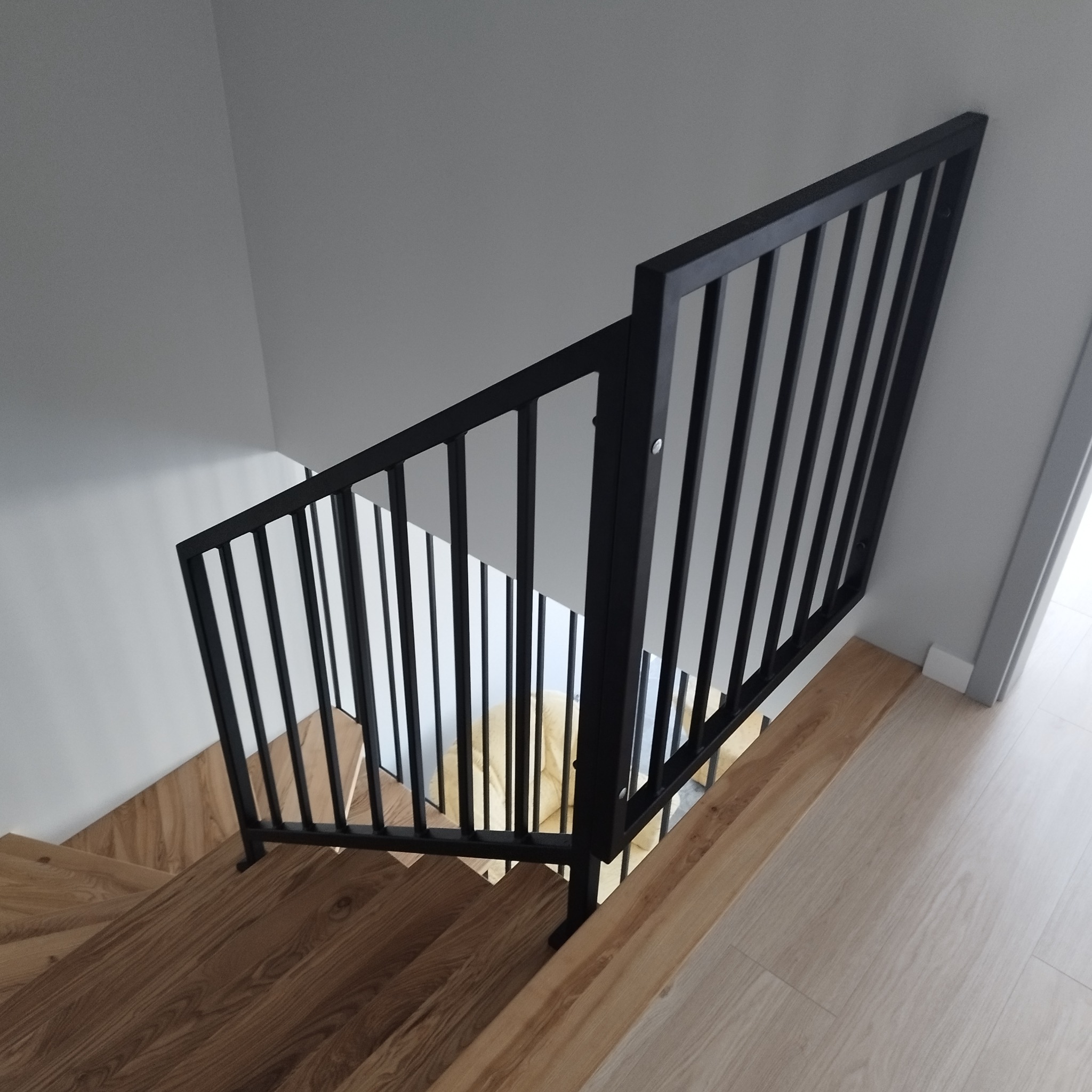 Nowoczesna, czarna balustrada metalowa z pionowymi szczeblami, zamontowana na drewnianych schodach w jasnym wnętrzu. Widok z góry, podkreślający minimalistyczny design i precyzję wykonania.