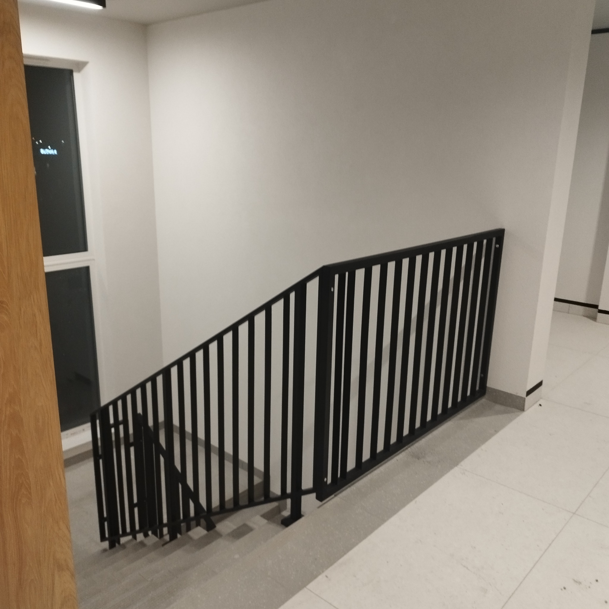 Minimalistyczne schody wewnętrzne z czarną metalową balustradą pionową, białe ściany i jasna podłoga z płytek.