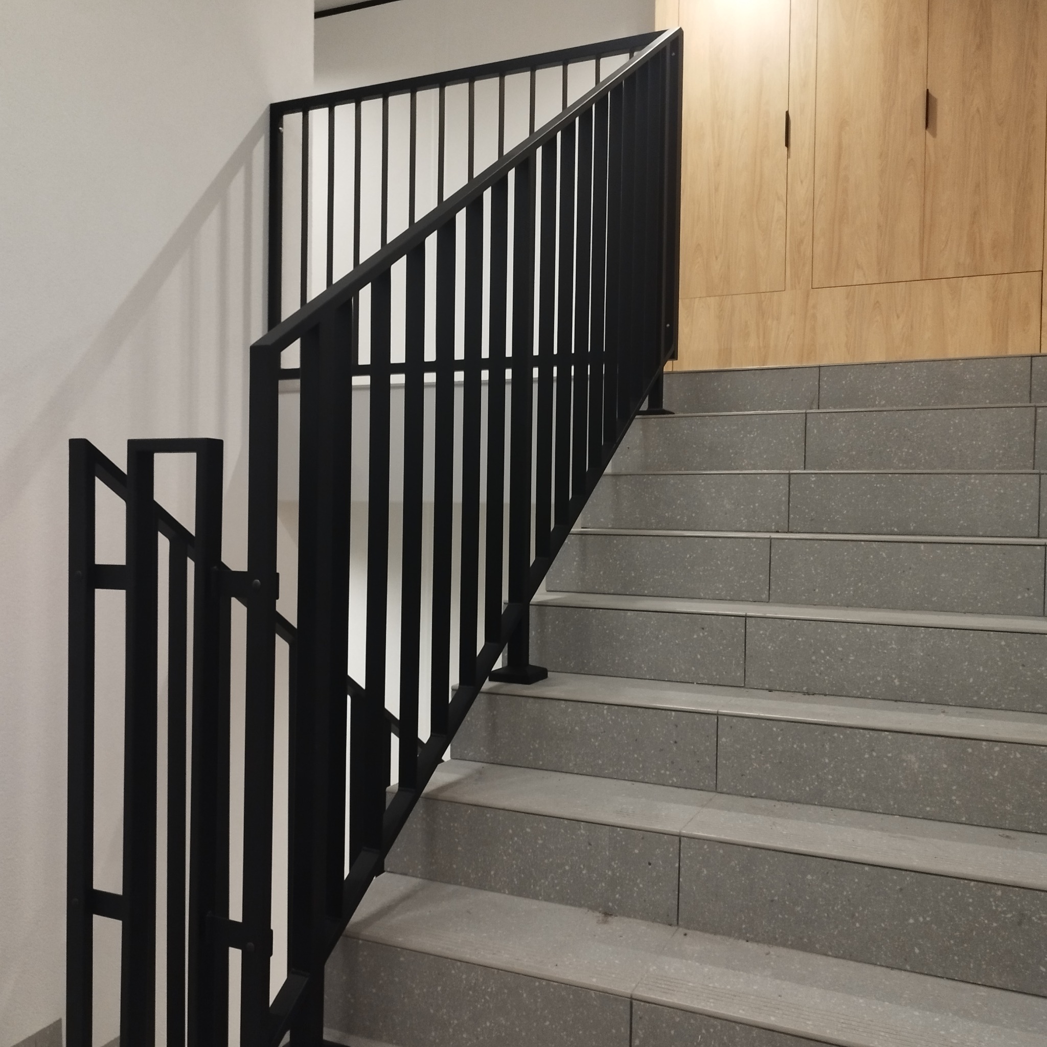 Nowoczesne schody z szarymi stopniami i czarną metalową balustradą o minimalistycznym designie, z drewnianą zabudową w tle.