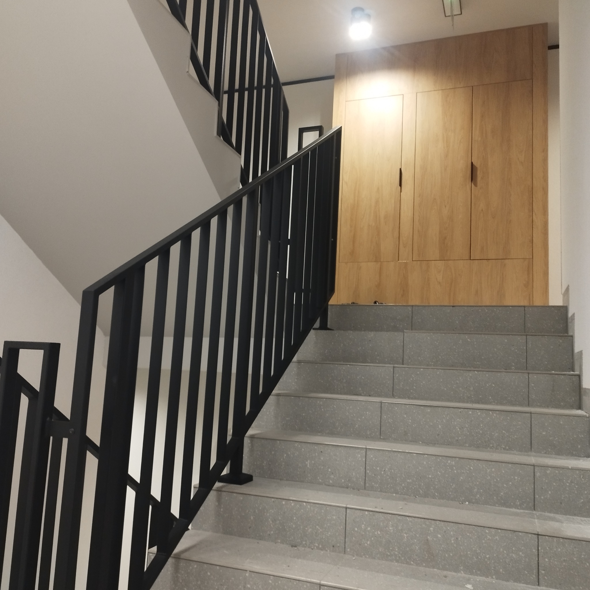 Wewnętrzne schody z szarych płytek z czarną metalową balustradą o pionowych szczeblach, obok drewnianej zabudowy ściennej.