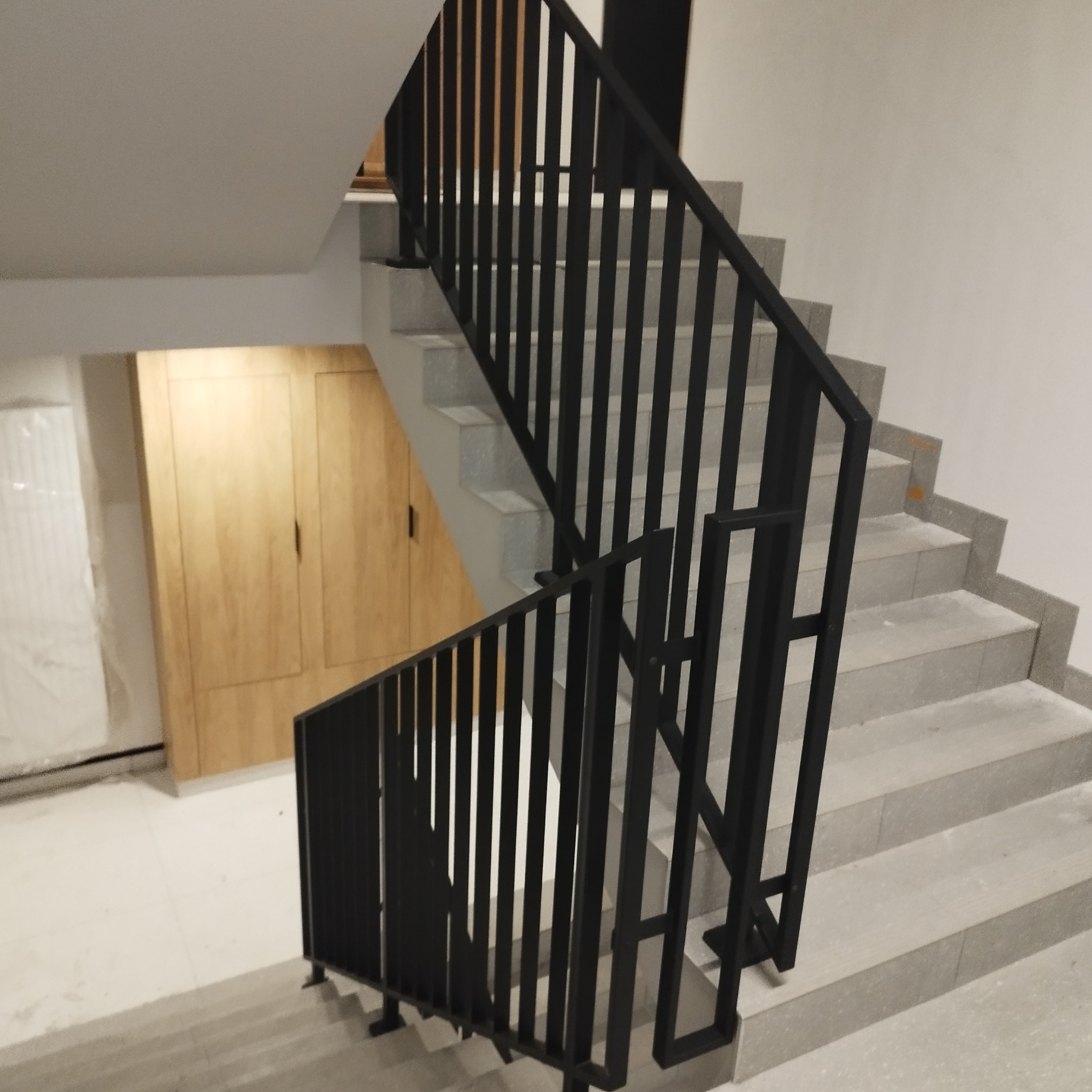 Wewnętrzne schody wykończone szarymi płytkami z czarną, metalową balustradą o pionowych prętach, widok z góry na dół, w tle drewniana zabudowa ściany.