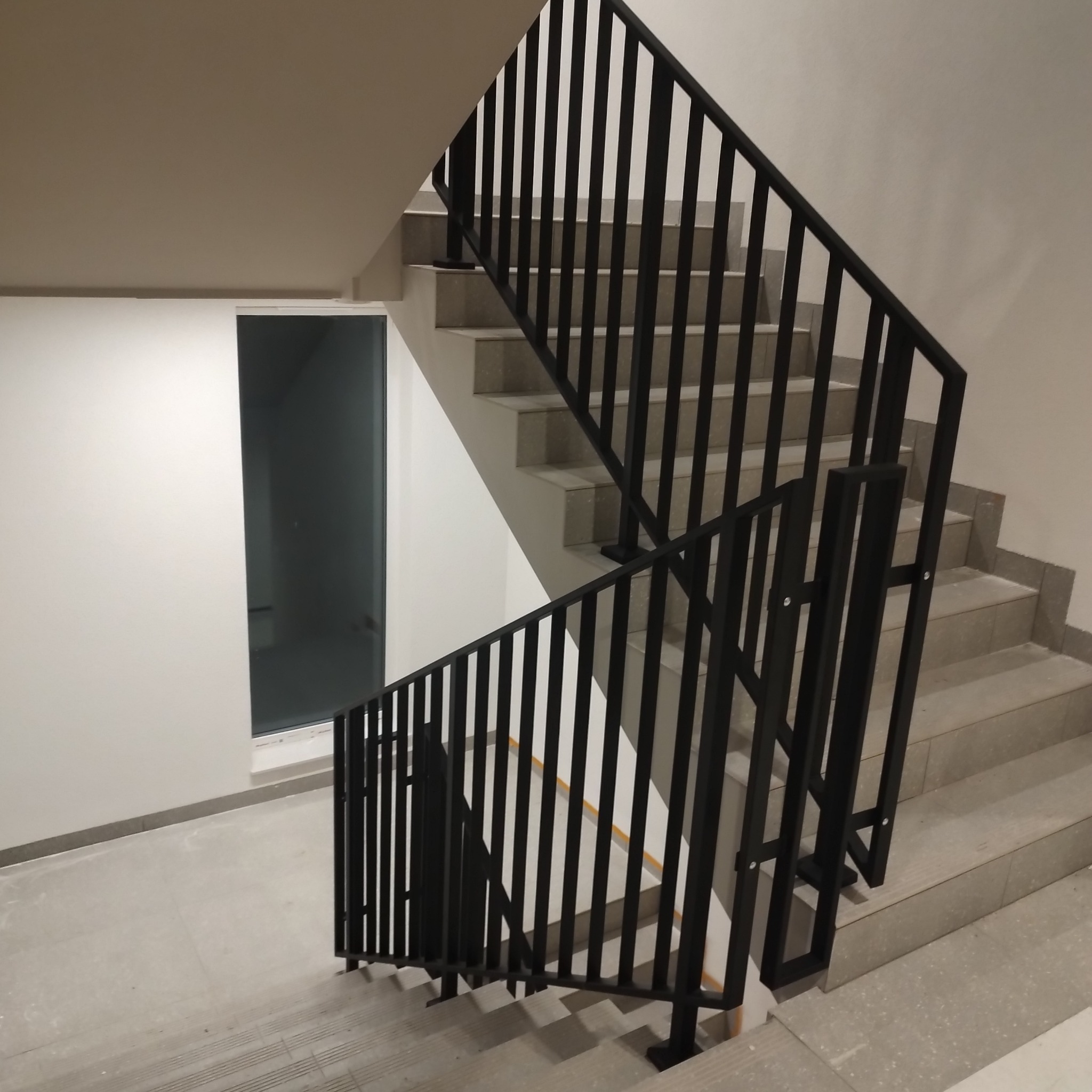 Wewnętrzne schody z szarymi stopniami i czarną metalową balustradą o pionowych szczeblach, widok z dołu na górę, neutralne oświetlenie, ściana z oknem w tle.