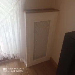 Biała zabudowa grzejnika z drewnianym blatem, ażurowy panel frontowy, zasłona w tle.