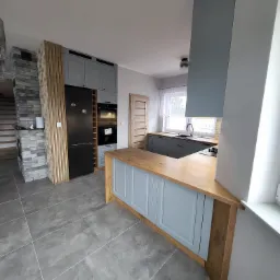 Nowoczesna szara kuchnia z drewnianym blatem, wyspą i akcentami w postaci drewnianych listew przy lodówce oraz ceglanego kominka w tle.