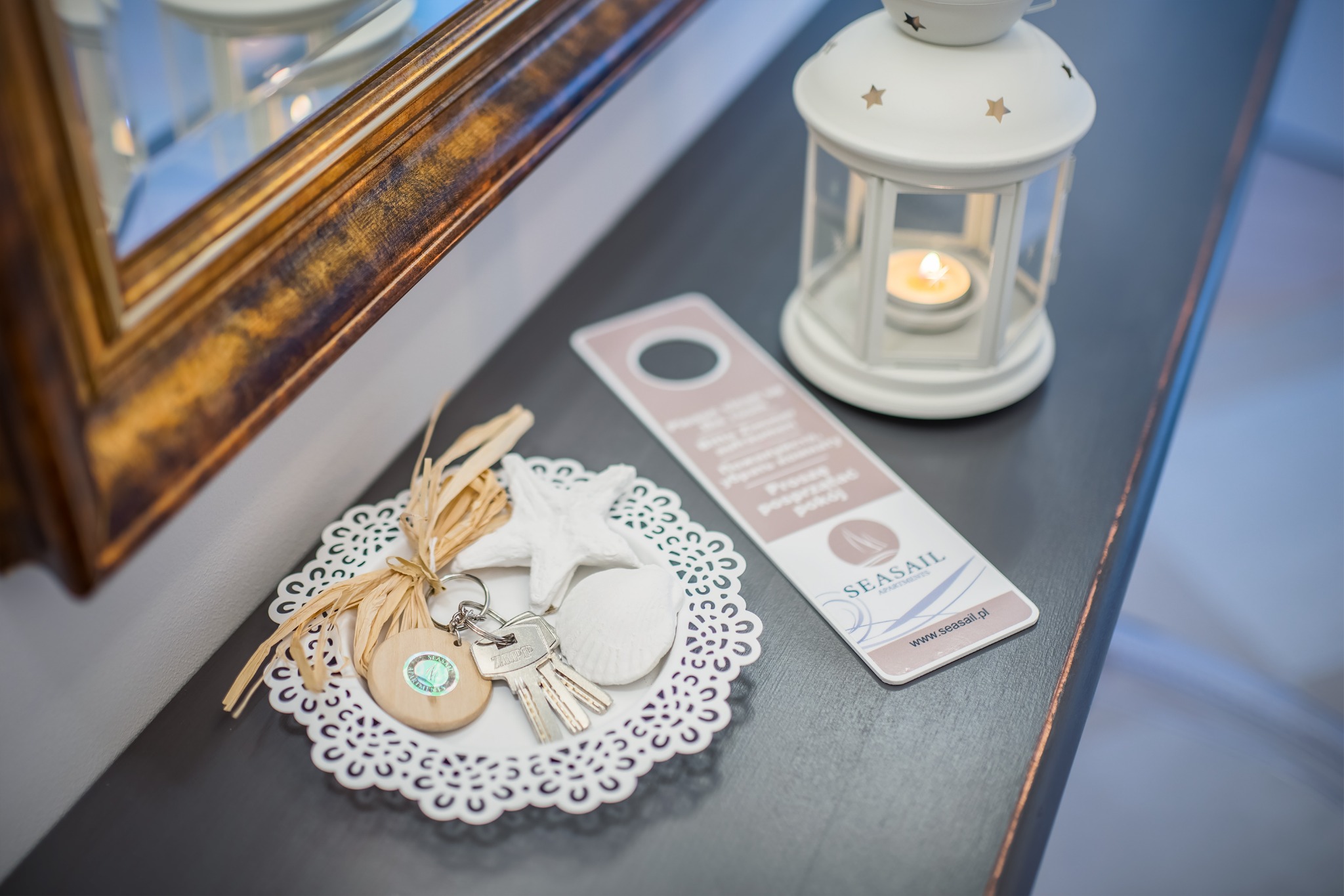Dekoracja w stylu nadmorskim na ciemnym blacie: lampion ze świecą, zawieszka na drzwi z logo Seasail Apartments, muszle, rozgwiazda i klucze na koronkowej serwetce obok lustra w złotej ramie.