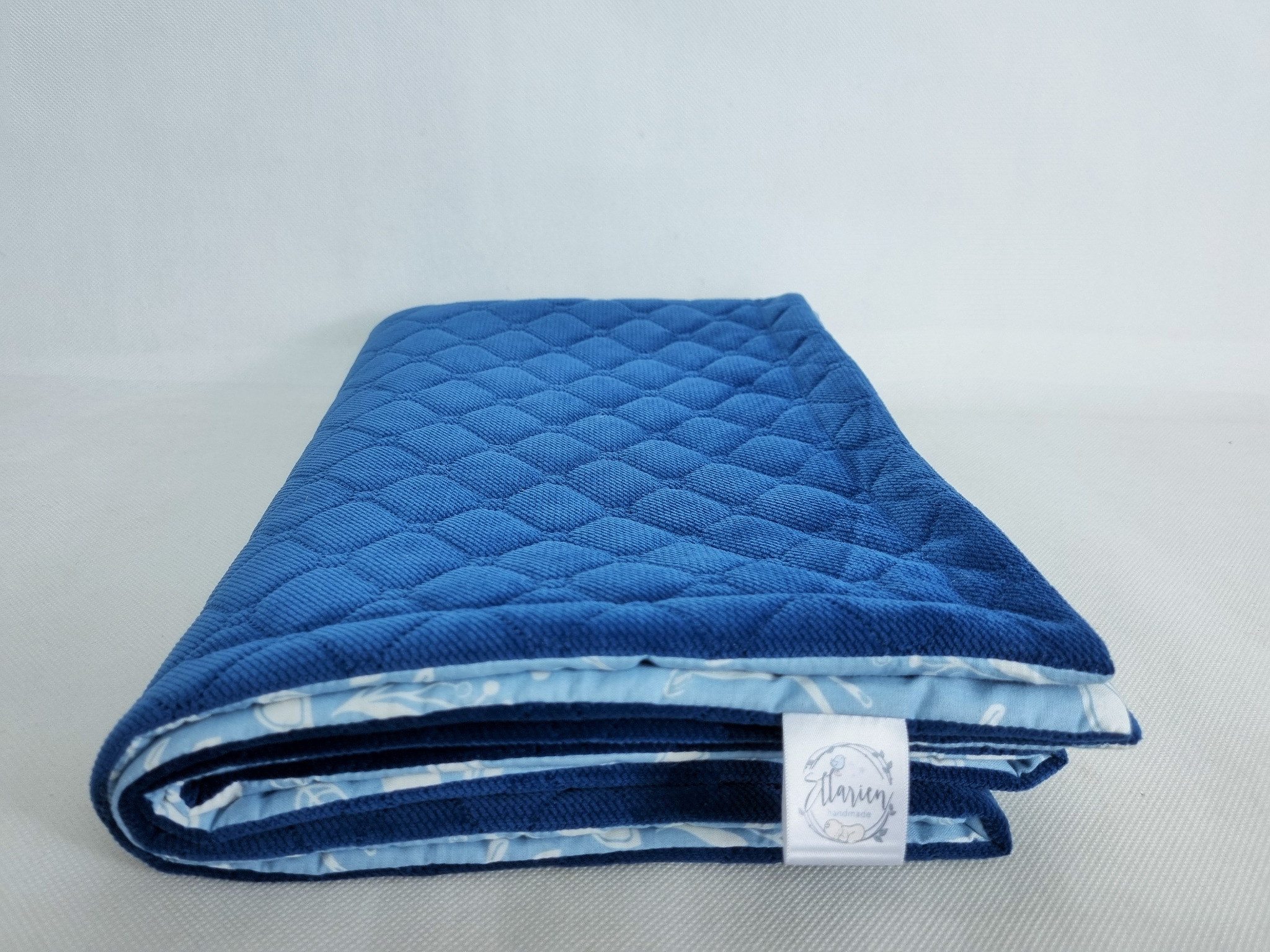 Złożona, pikowana kołderka dziecięca w odcieniach niebieskiego z dekoracyjną, jasną podszewką z wzorem i wszytą metką z logo producenta.