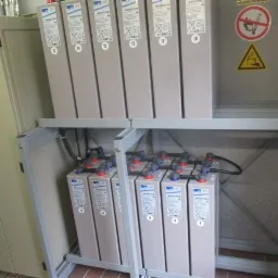 Baterie akumulatorowe w szafie zasilającej, widoczny panel sterujący i znaki ostrzegawcze na ścianie, instalacja w pomieszczeniu technicznym.