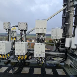 Rozbudowana instalacja telekomunikacyjna na dachu budynku z widokiem na okolicę, liczne moduły i okablowanie, szara metalowa konstrukcja wsporcza.