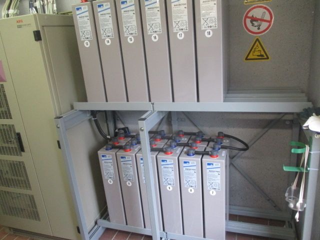 Baterie akumulatorowe w szafie zasilającej, widoczny panel sterujący i znaki ostrzegawcze na ścianie, instalacja w pomieszczeniu technicznym.