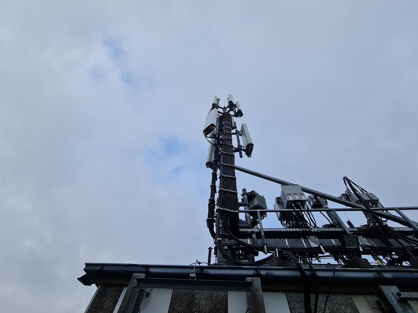Maszt telekomunikacyjny z antenami i okablowaniem, widoczny z dołu na tle pochmurnego nieba.
