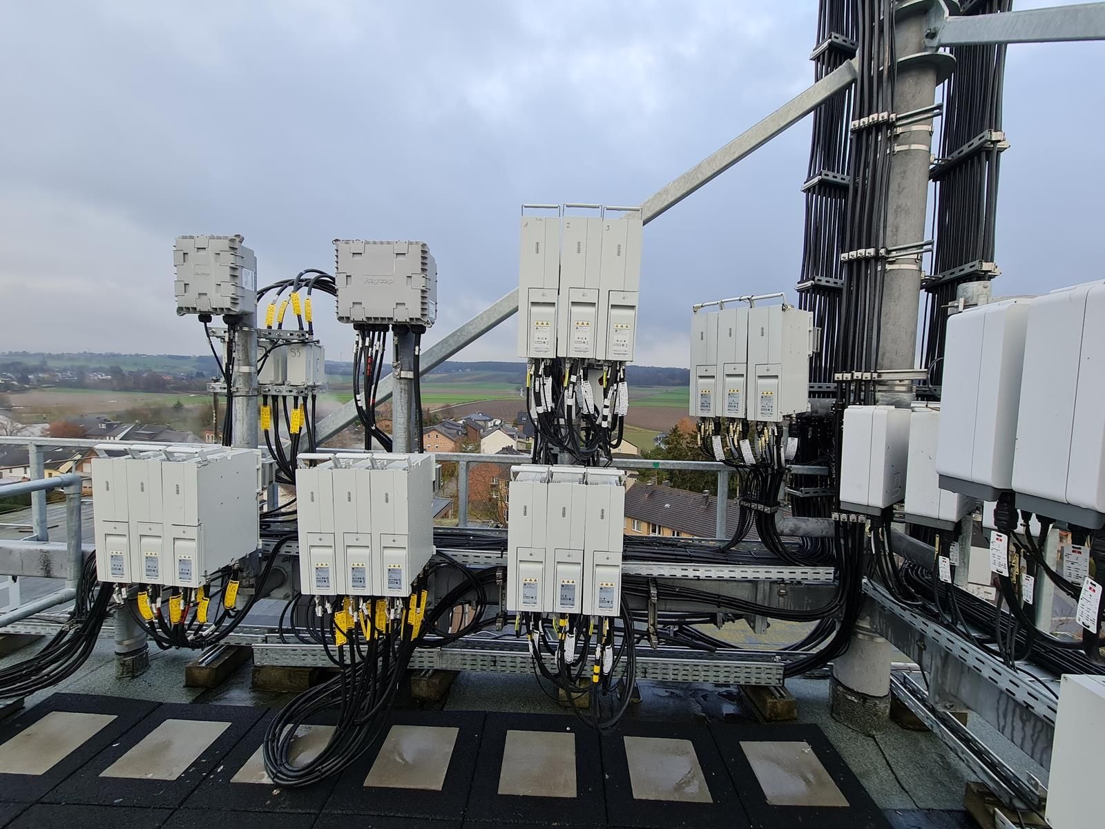 Rozbudowana instalacja telekomunikacyjna na dachu budynku z widokiem na okolicę, liczne moduły i okablowanie, szara metalowa konstrukcja wsporcza.