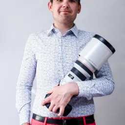 Uśmiechnięty fotograf trzymający duży obiektyw Canon EF 300mm f/2.8L IS II USM, ubrany w wzorzystą koszulę i czerwone spodnie.