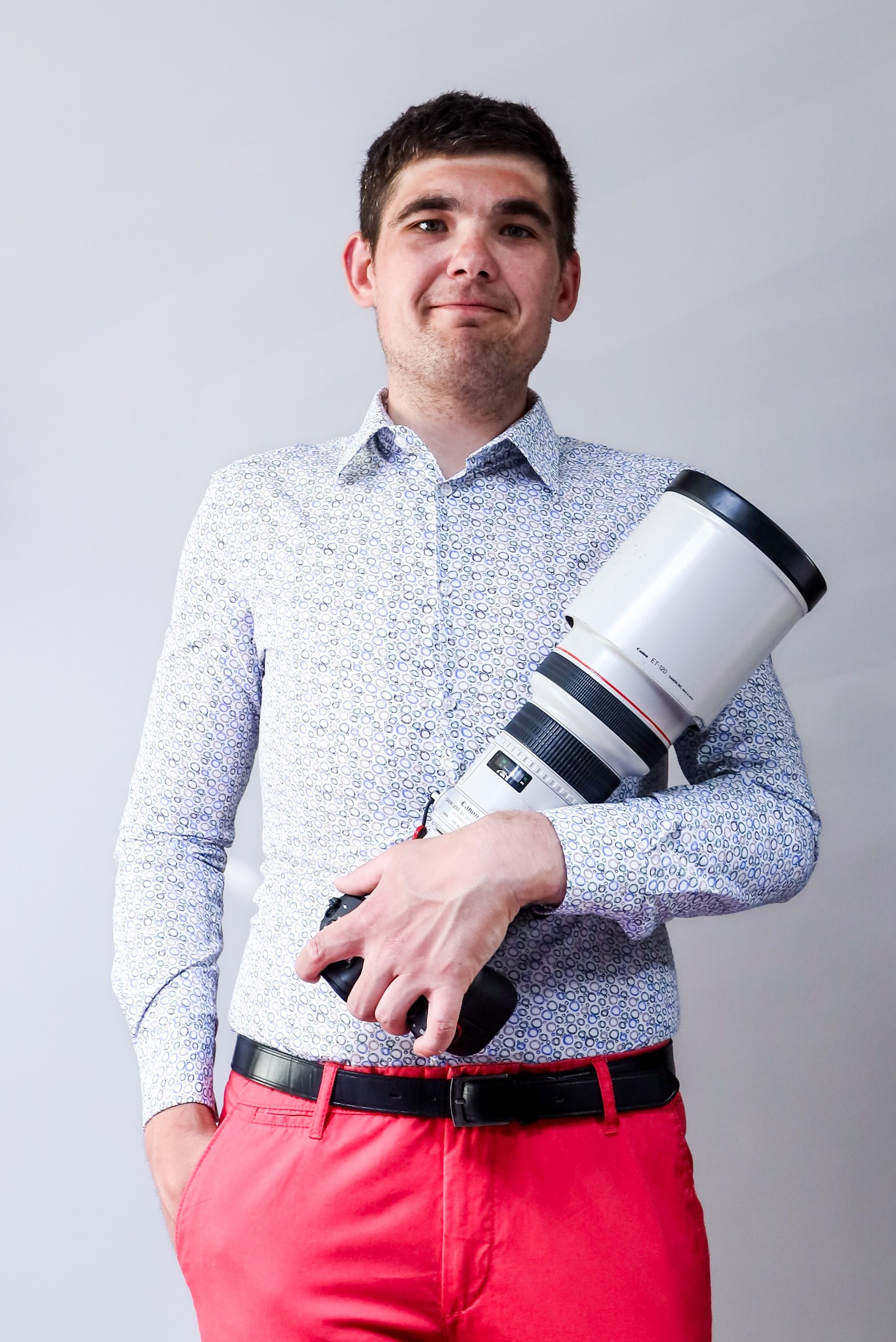 Uśmiechnięty fotograf trzymający duży obiektyw Canon EF 300mm f/2.8L IS II USM, ubrany w wzorzystą koszulę i czerwone spodnie.