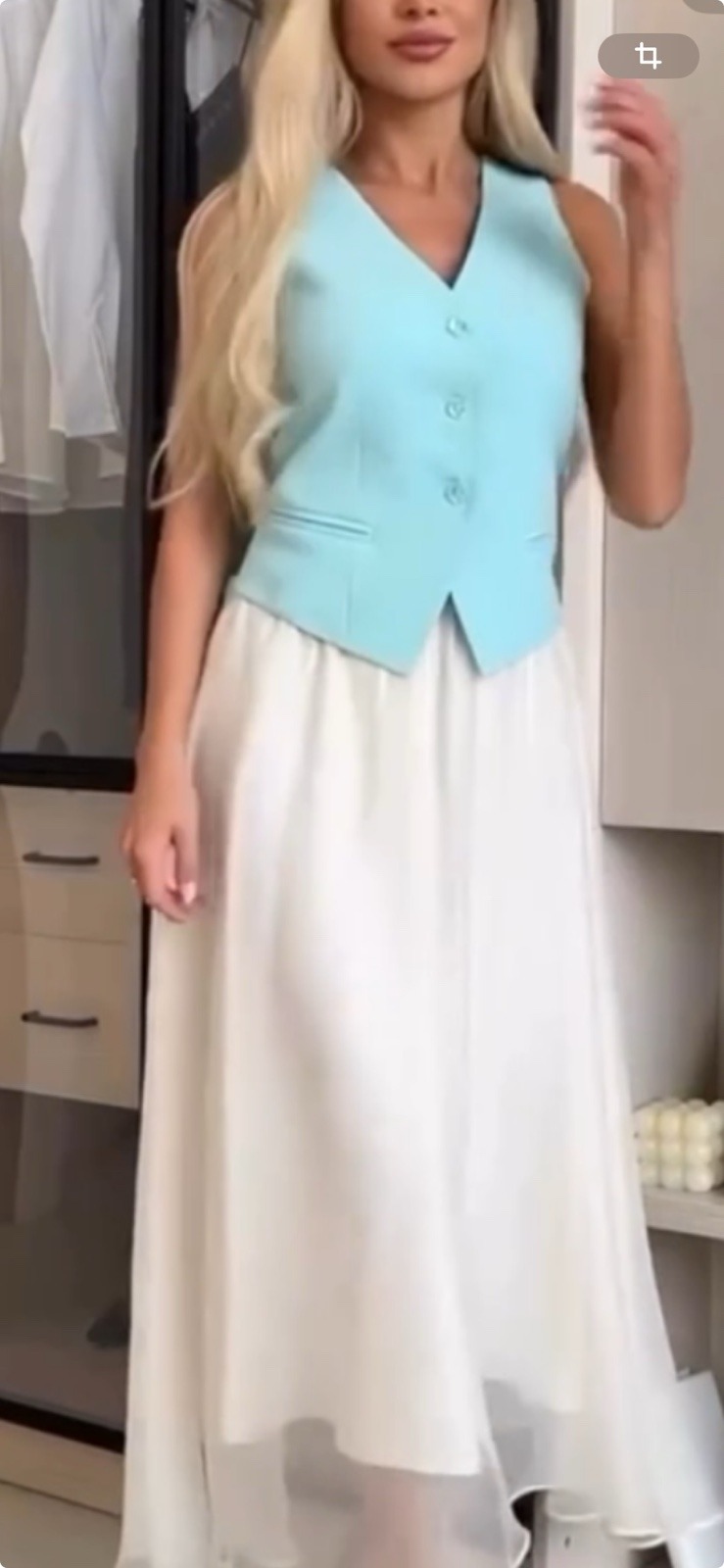 Kobieta prezentuje stylizację z turkusową kamizelką zapinaną na trzy guziki i długą, zwiewną, białą spódnicą, na tle szafy z przeszklonymi drzwiami.
