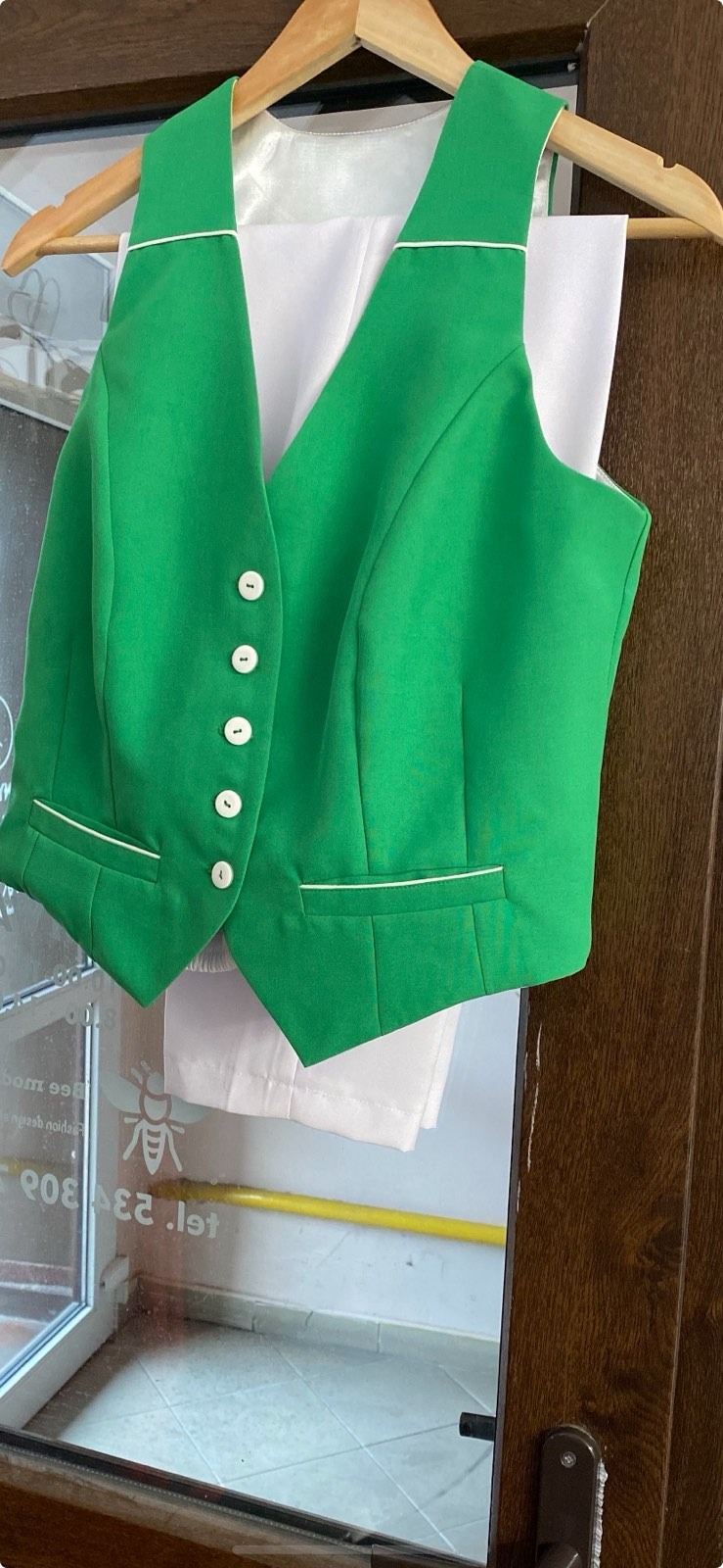 Jasnozielona kamizelka z białymi guzikami i wykończeniem, zawieszona na drewnianym wieszaku, z białymi spodniami w tle, widoczne odbicie okna i fragment drzwi.