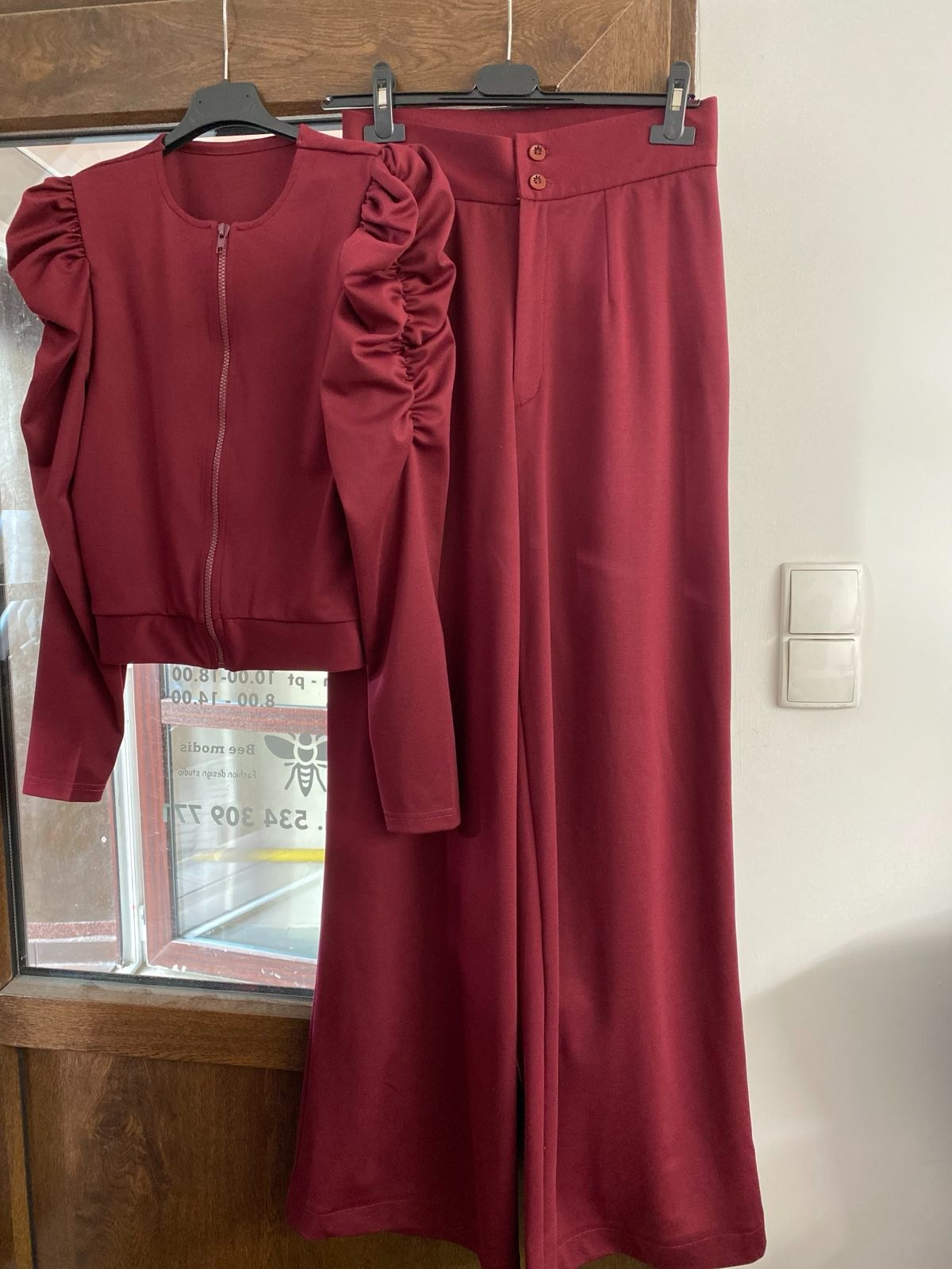 Komplet damski w kolorze burgundowym: bluza zapinana na zamek z bufkami na ramionach i szerokie spodnie z wysokim stanem, wiszące na wieszaku przed oknem.