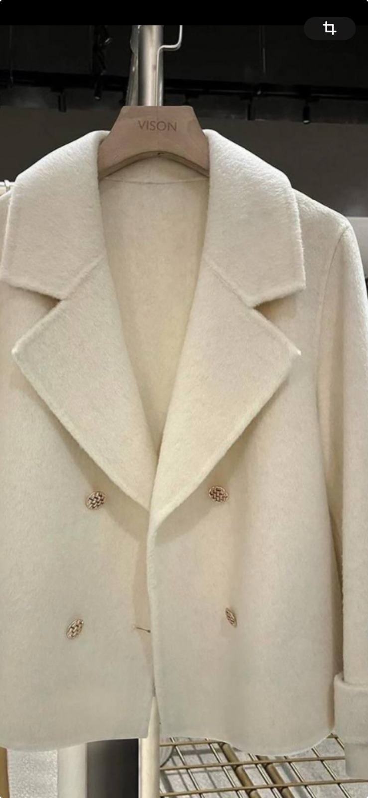 Krótki, kremowy płaszcz damski z ozdobnymi guzikami, wiszący na wieszaku marki VISON.