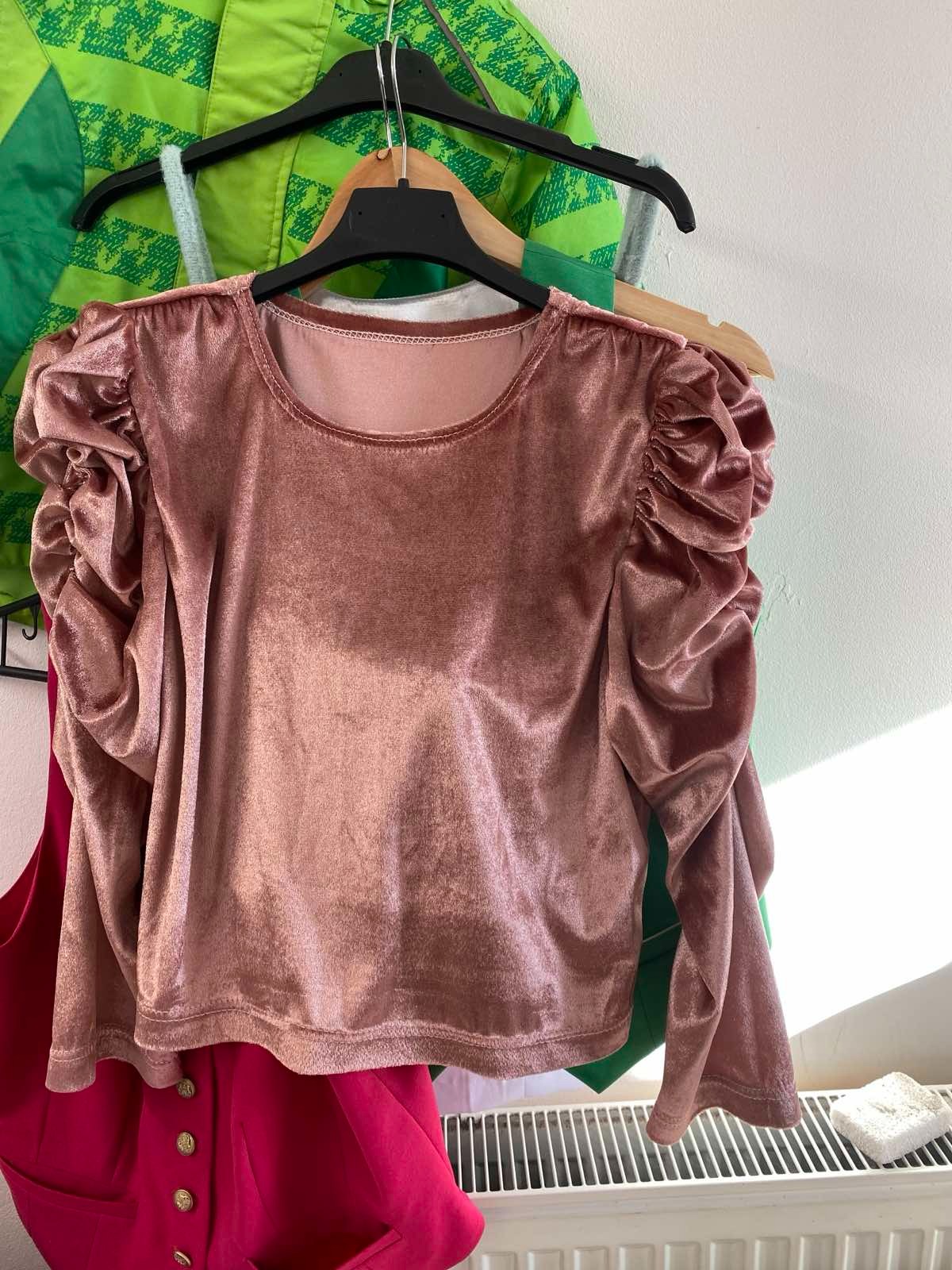 Bluzka z różowego weluru z bufowanymi rękawami, wisząca na wieszaku, z widocznymi innymi ubraniami w tle, w tym zielona kurtka i różowa kamizelka z guzikami.