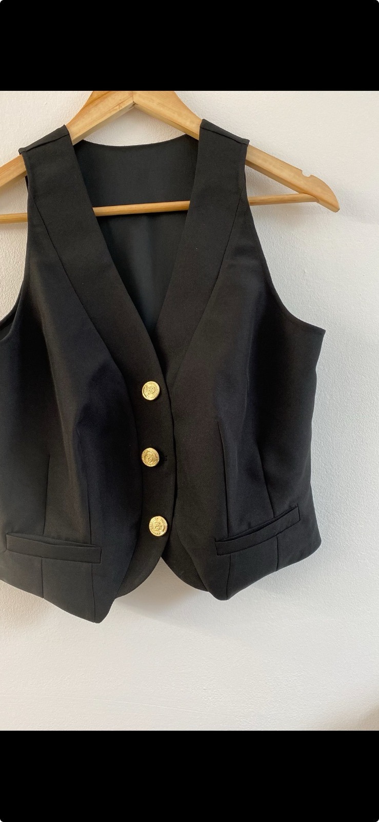 Czarna, krótka kamizelka damska z trzema złotymi guzikami, wisząca na drewnianym wieszaku na tle białej ściany.