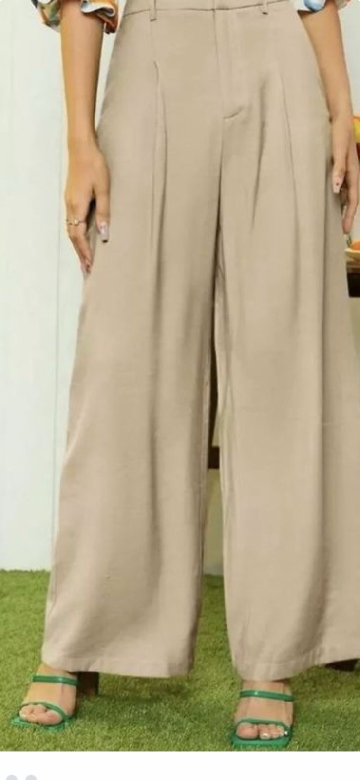 Beżowe, szerokie spodnie damskie z zakładkami, prezentowane na modelce z zielonymi sandałami na sztucznej trawie.