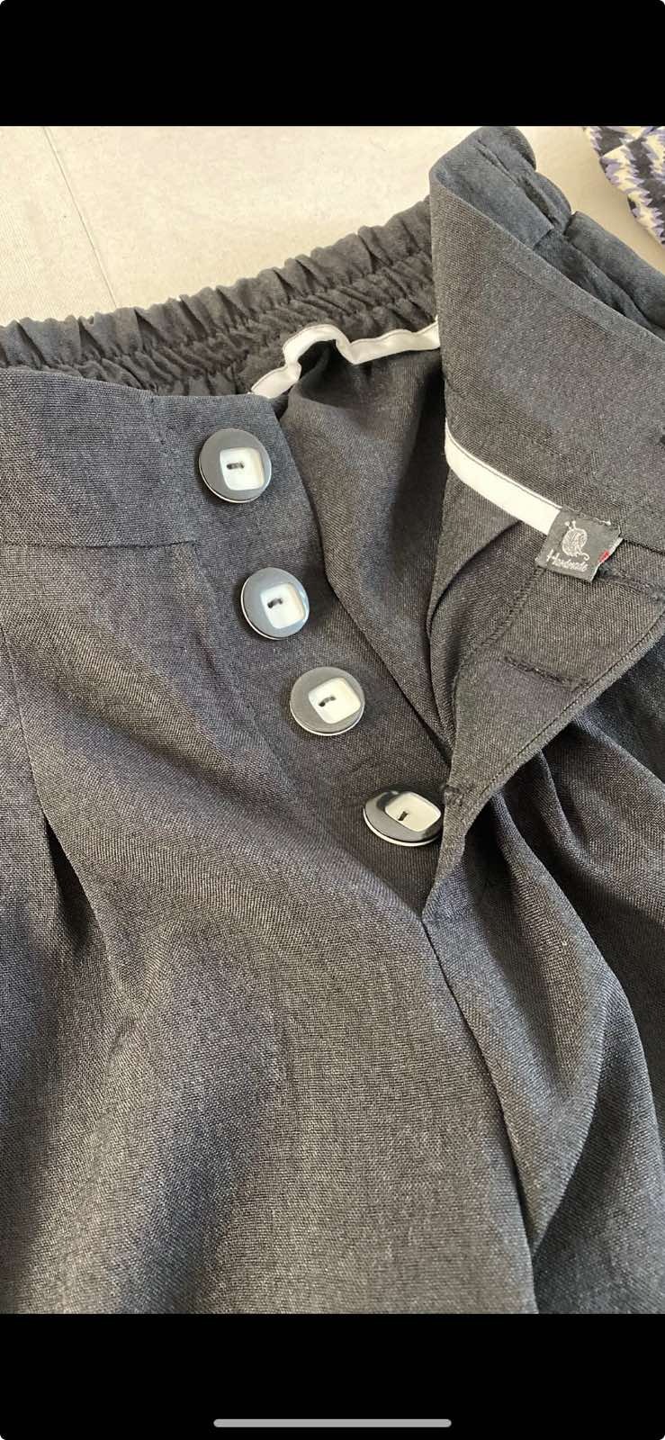 Szare spodnie damskie z wysokim stanem, zapinane na cztery duże guziki z szarą obwódką i białym środkiem, widoczna metka z napisem 'Handmade'.