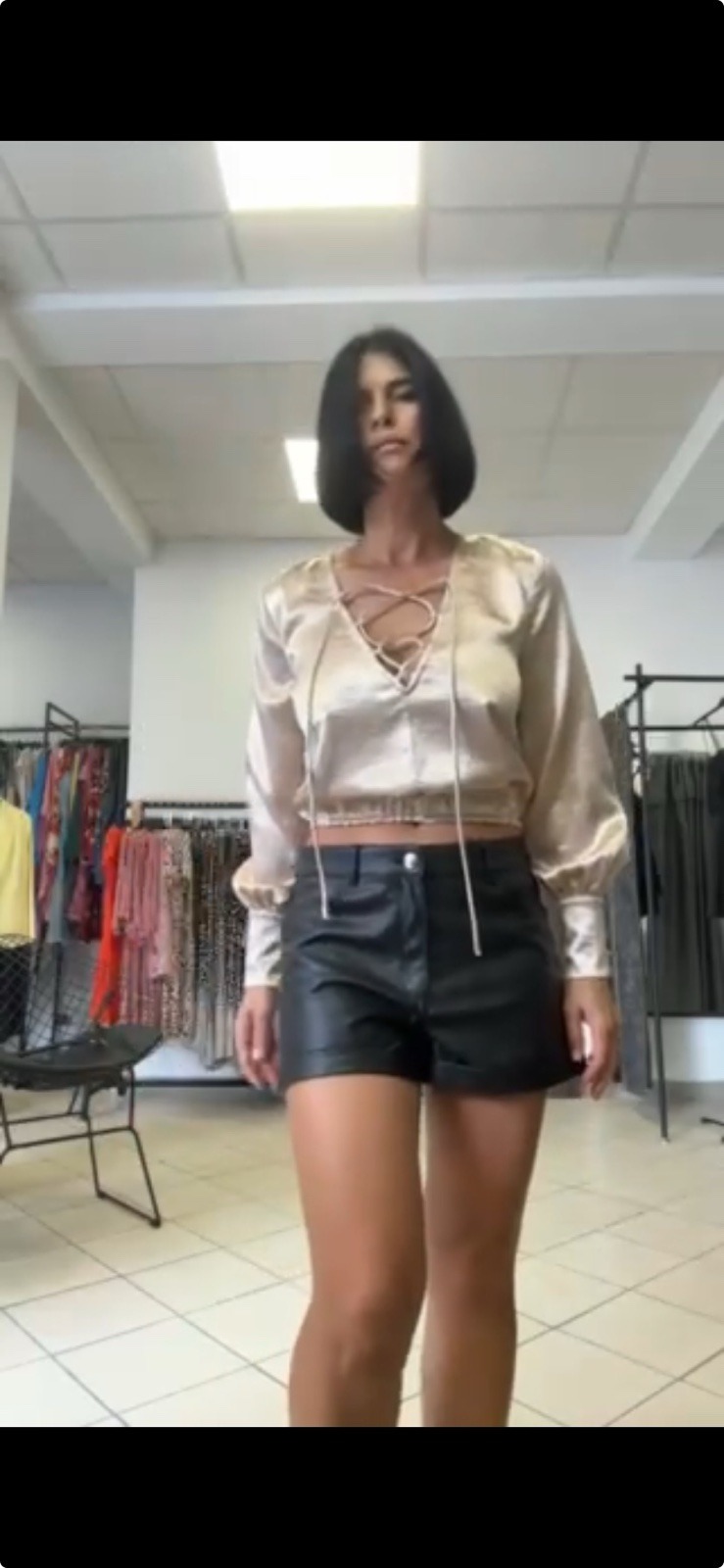 Kobieta w sklepie odzieżowym prezentuje stylizację: satynowa bluzka z wiązaniem na dekolcie i czarne skórzane szorty.