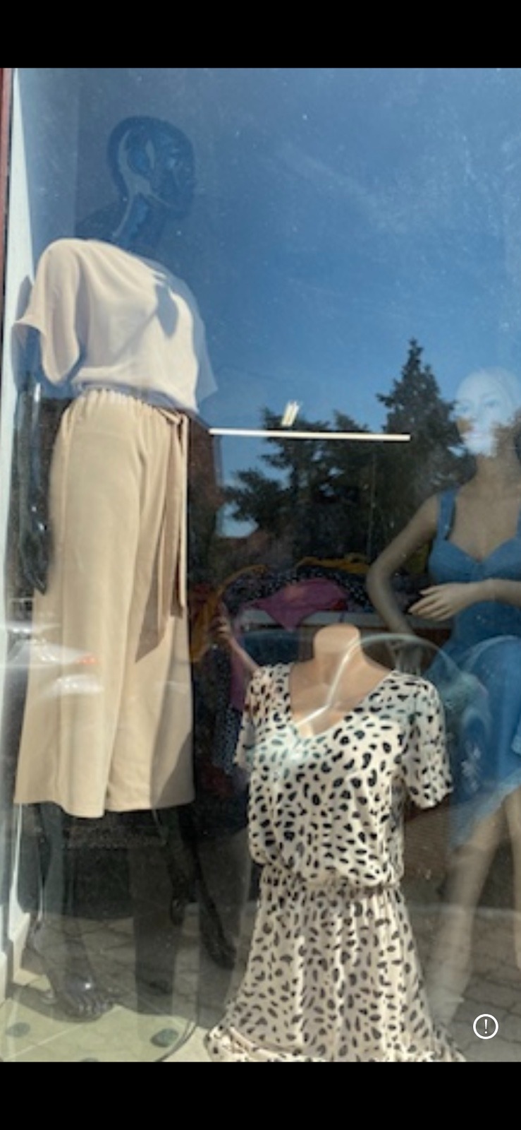 Witryna sklepowa z trzema manekinami prezentującymi odzież damską: beżowy komplet z bluzką i spodniami, sukienka w panterkę oraz niebieska sukienka na ramiączkach.