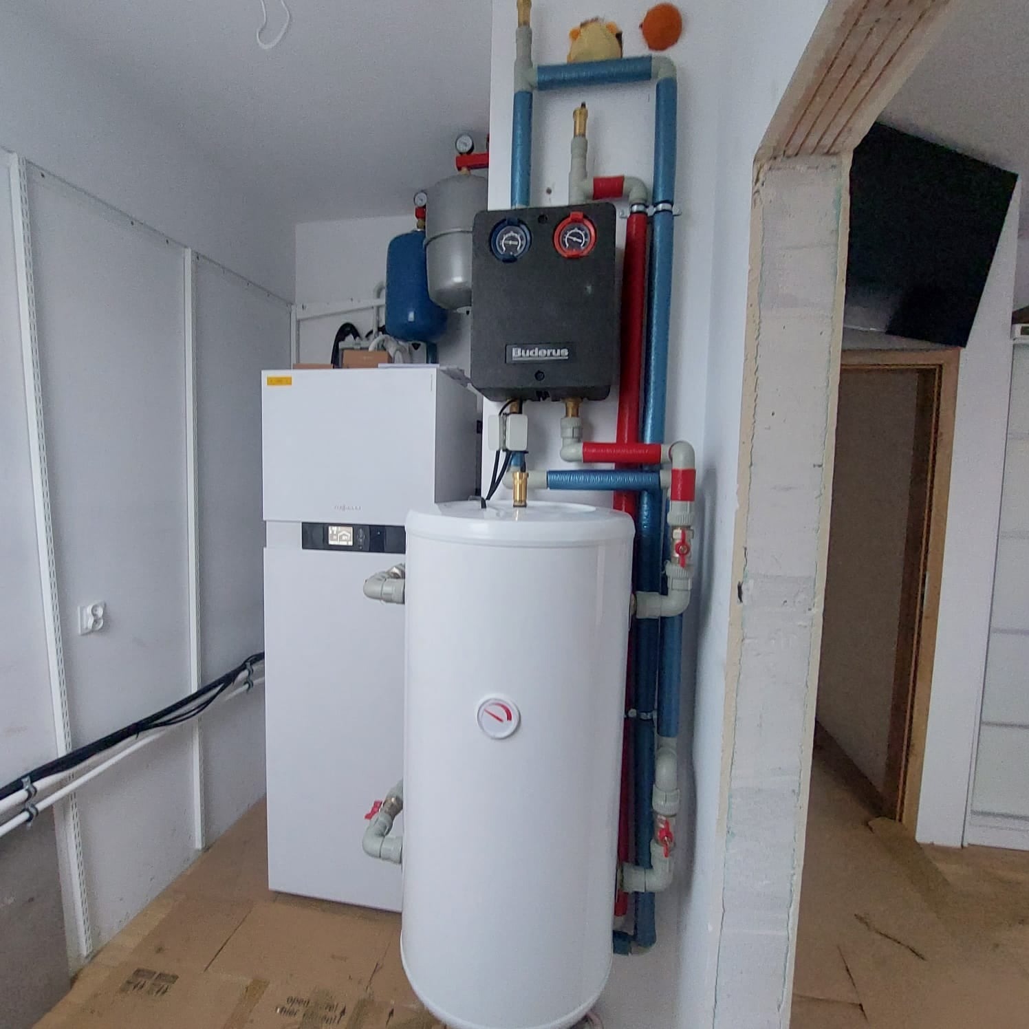 Biała instalacja grzewcza Buderus z zbiornikiem i rurami w pomieszczeniu gospodarczym, widoczne elementy sterujące i zabezpieczające, pluszowa zabawka na rurze