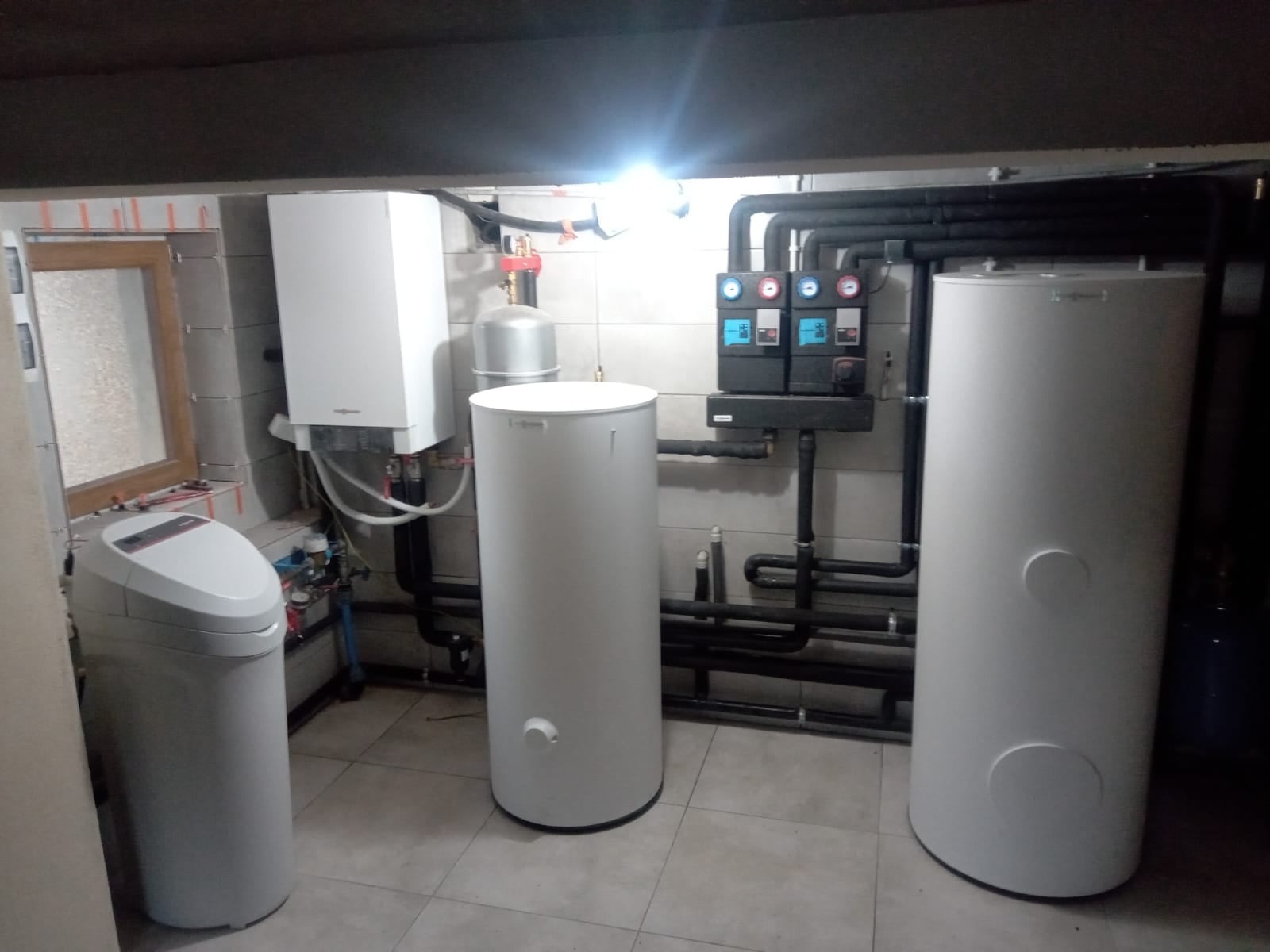 Instalacja w kotłowni: biały zasobnik ciepłej wody, wiszący piec grzewczy i system rur z manometrami na kafelkowanej ścianie.