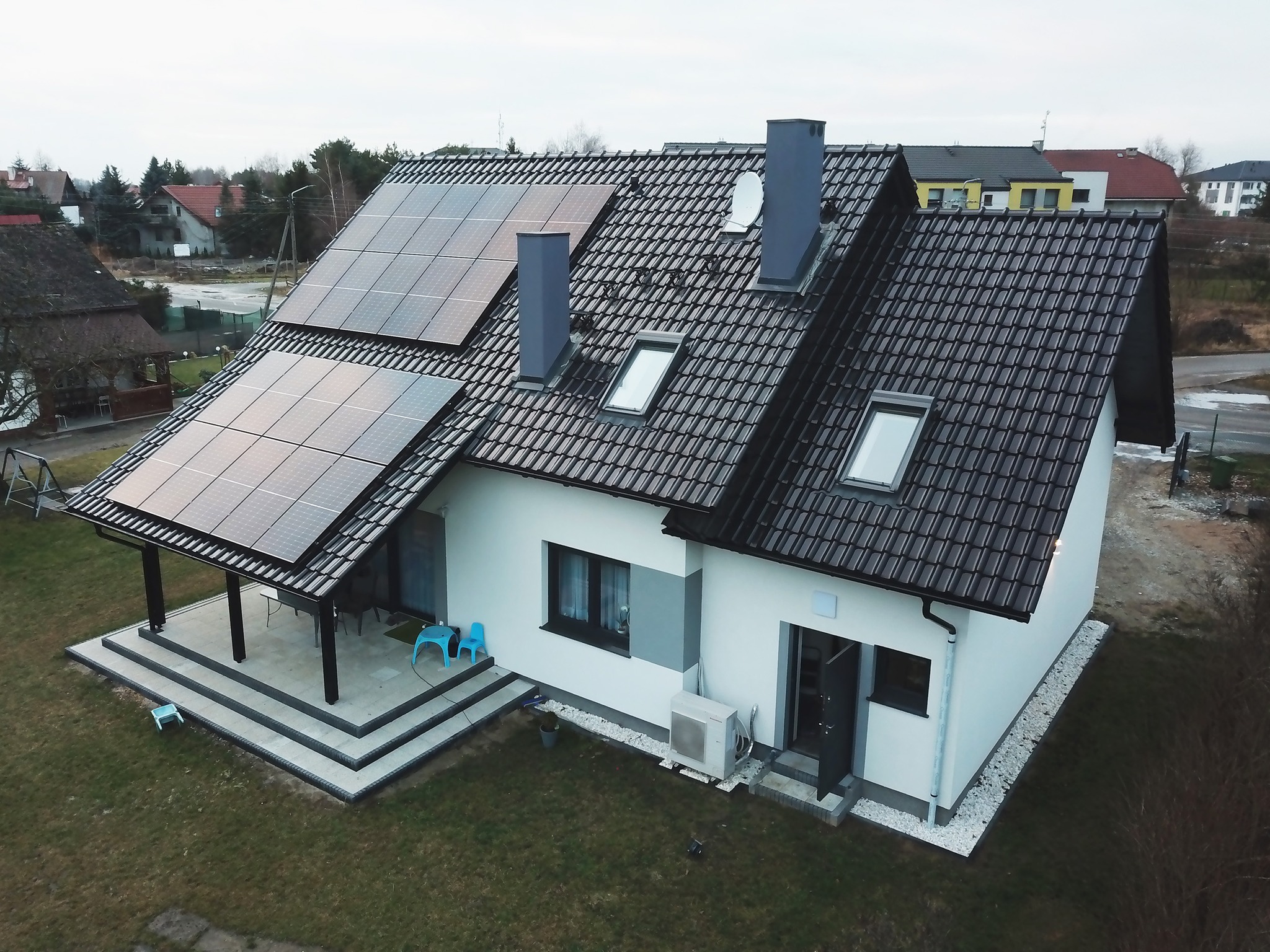Nowoczesny dom jednorodzinny z panelami fotowoltaicznymi na dachu, widok z góry, z klimatyzatorem na zewnątrz i trzema niebieskimi krzesłami na tarasie.