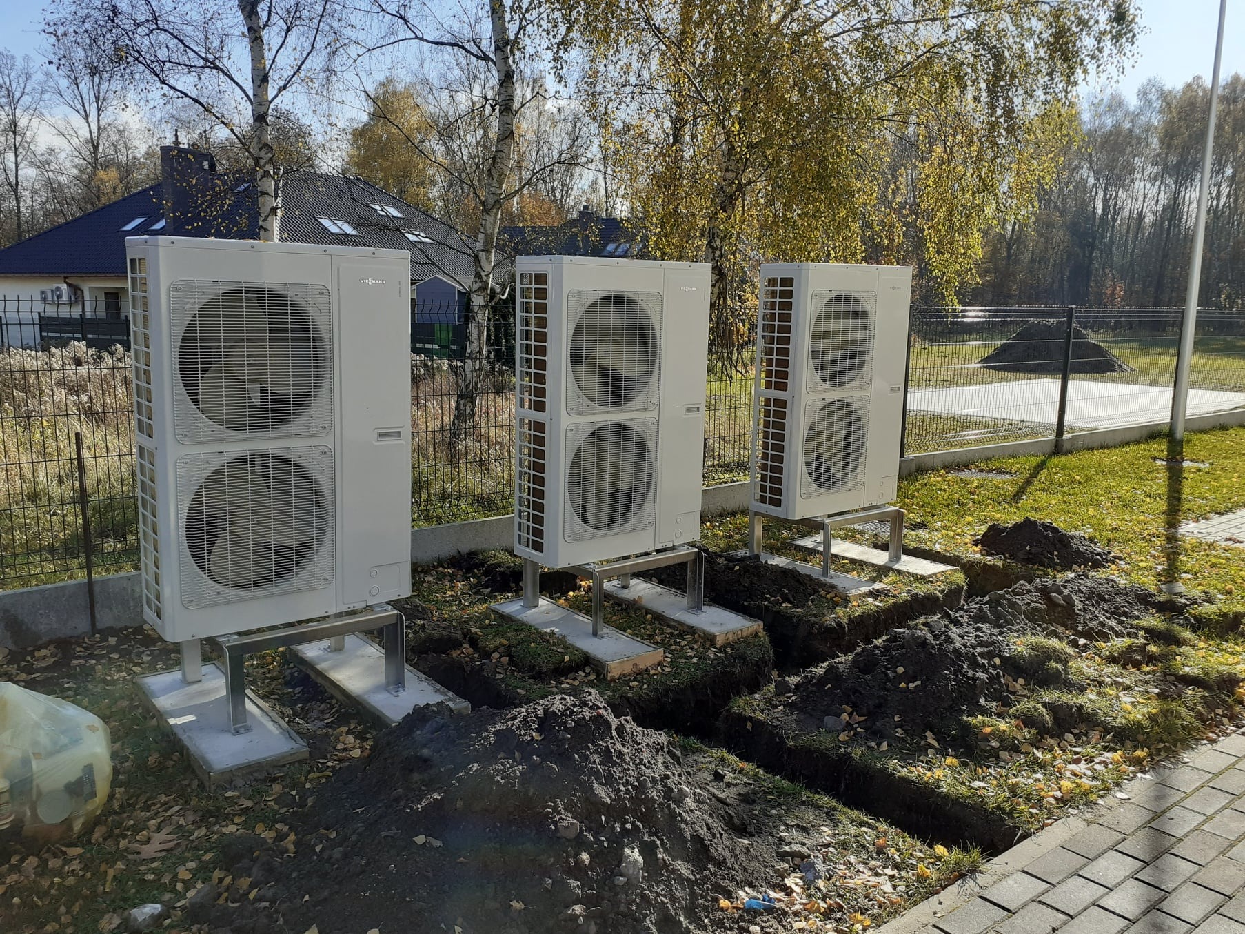 Trzy zewnętrzne jednostki pompy ciepła Viessmann zamontowane na betonowych podstawach i metalowych konstrukcjach, z wykopanymi rowami w ziemi, liście na trawie, w tle dom z ciemnym dachem i drzewa.