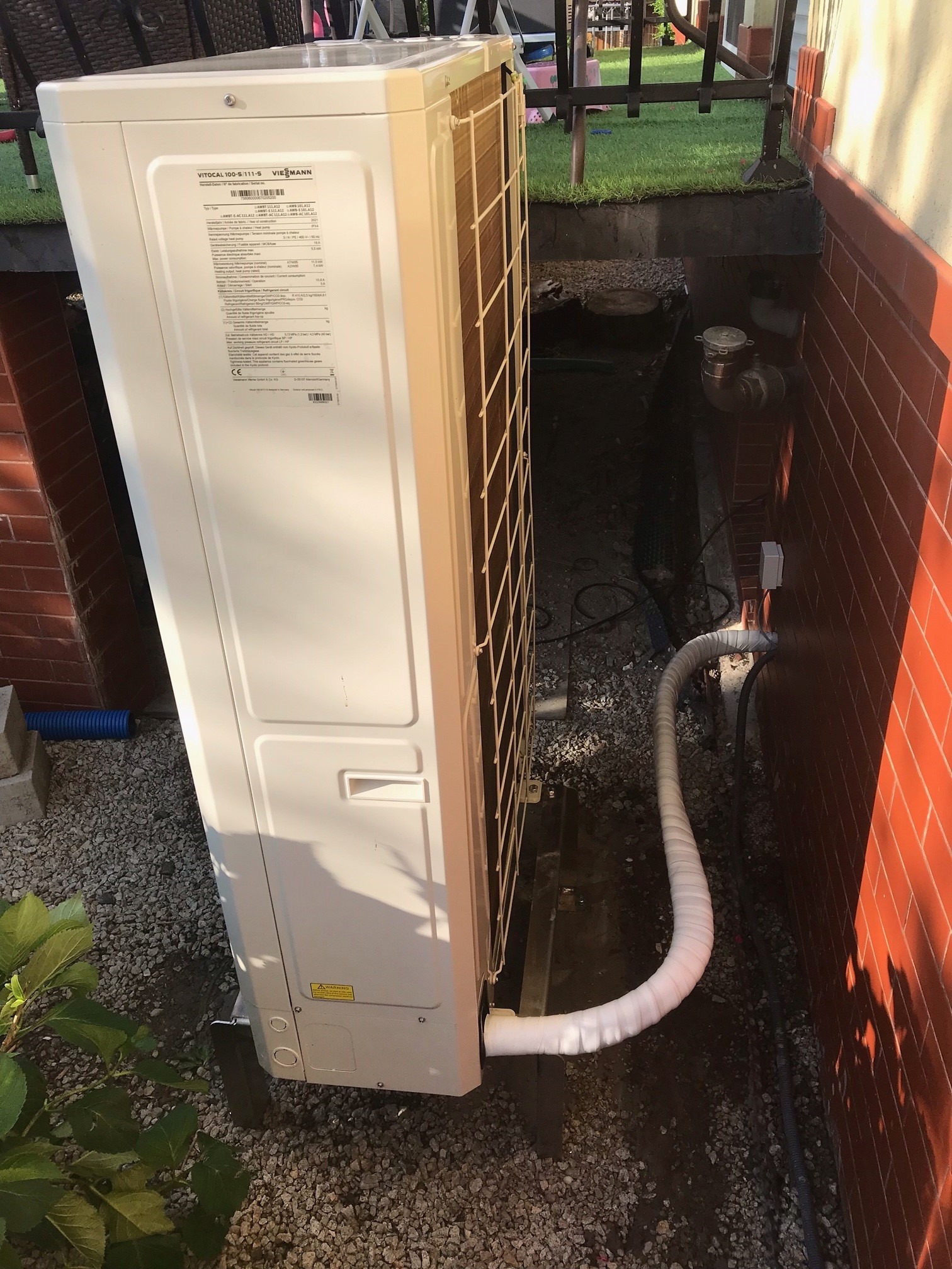 Zewnętrzna jednostka pompy ciepła Viessmann Vitocal 100-S/111-S, zainstalowana przy ceglanej ścianie budynku, z widocznymi rurami odprowadzającymi skropliny w izolacji, otoczona żwirem i zielenią.