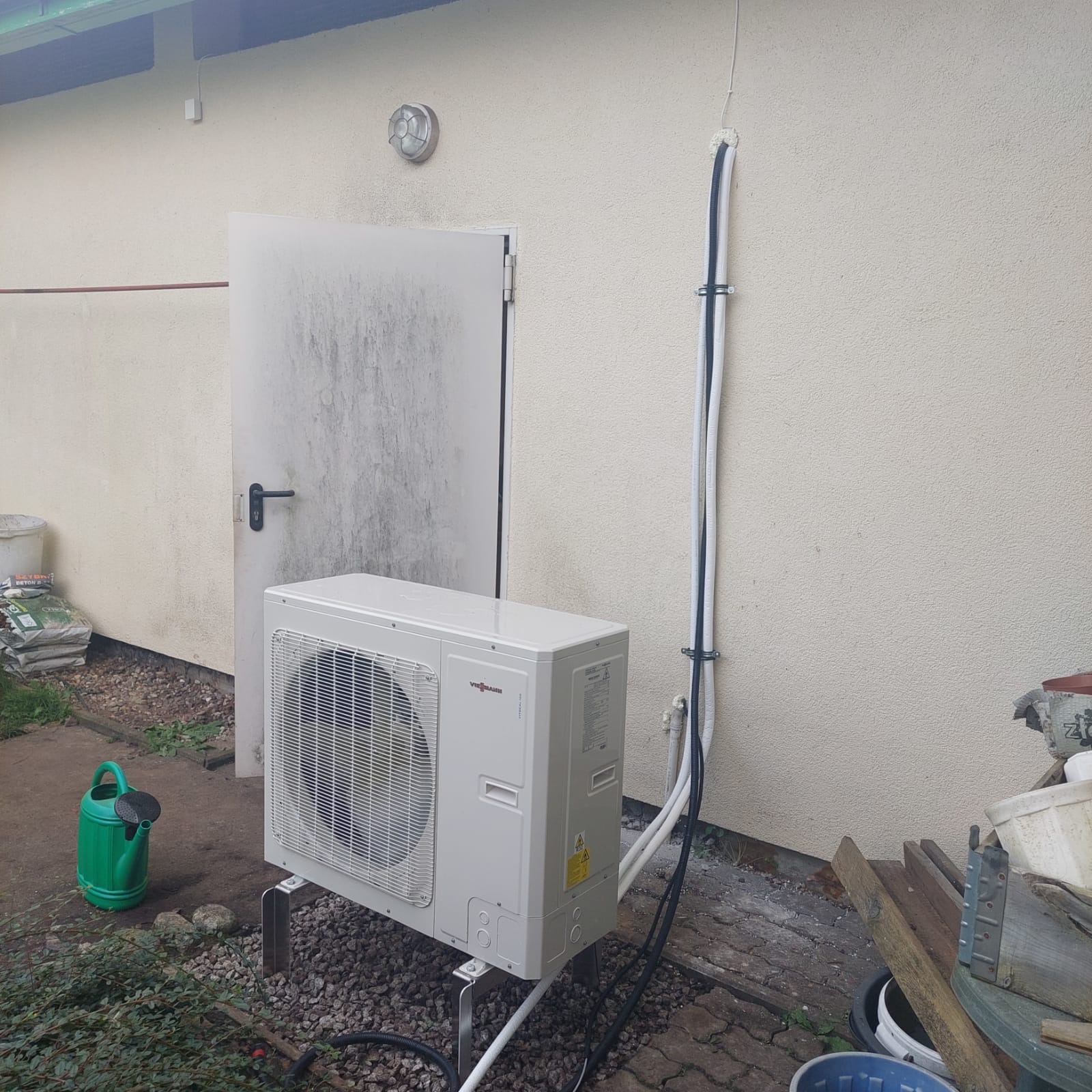 Zewnętrzna jednostka grzewcza Viessmann zamontowana na metalowych wspornikach, z widocznymi rurami przymocowanymi do ściany budynku, obok znajduje się zielona konewka.