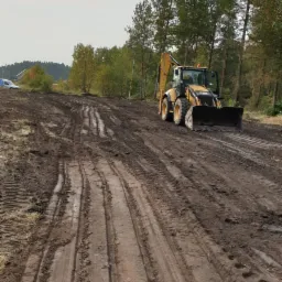 Żółta koparko-ładowarka Caterpillar na placu budowy po deszczu, widoczne głębokie ślady opon w błotnistym gruncie, w tle las i fragment białego samochodu dostawczego.