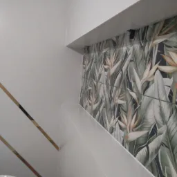 Ściana łazienki wyłożona białymi płytkami z dekoracyjnymi złotymi paskami oraz płytkami z motywem tropikalnych liści i kwiatów strelicji.