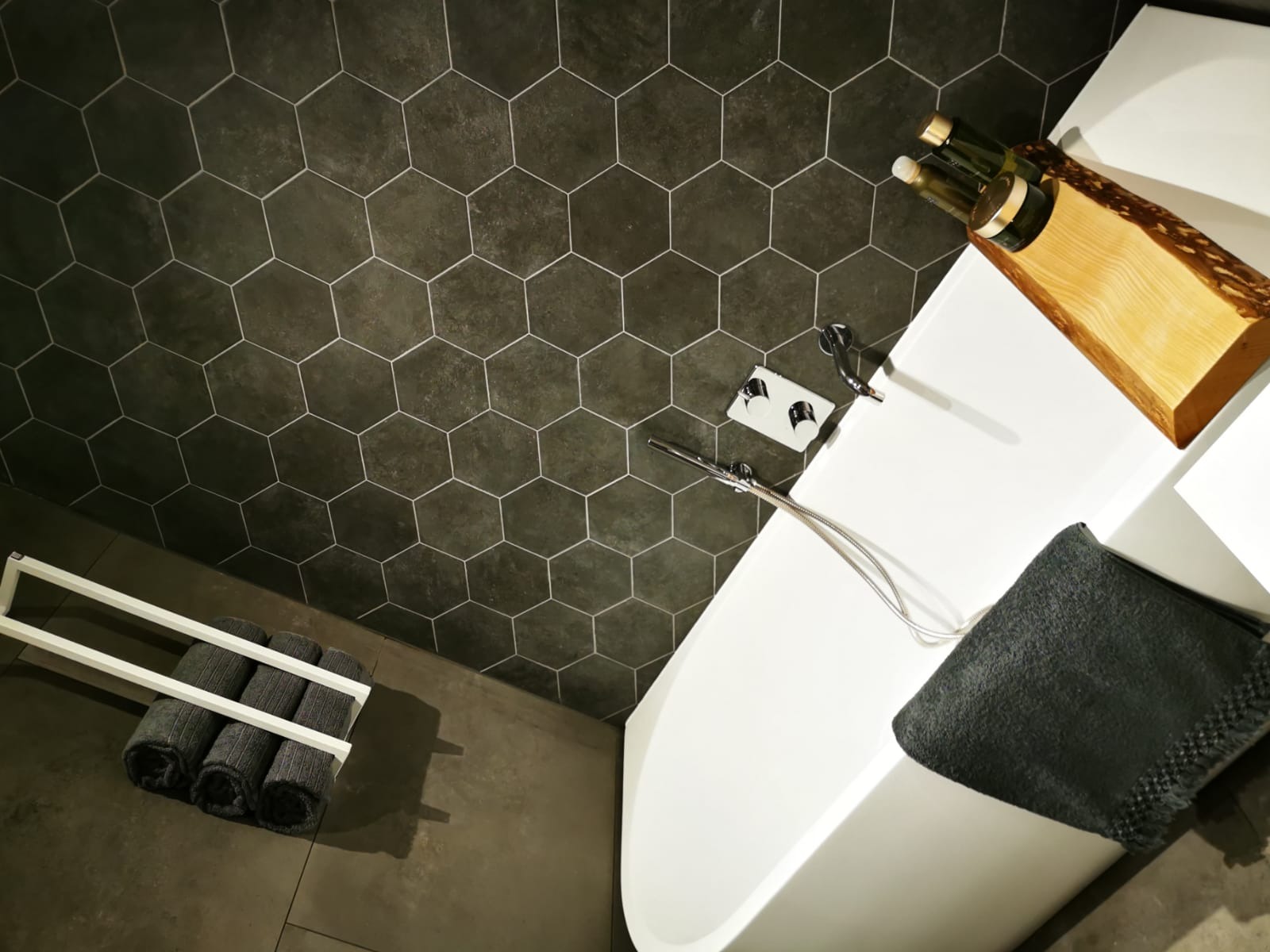 Nowoczesna łazienka z heksagonalnymi płytkami w odcieniach szarości, białą wanną wolnostojącą, drewnianym organizerem na kosmetyki i ręcznikami na wieszaku, widziana z góry.