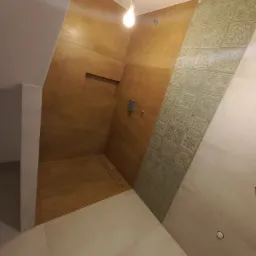 Wnętrze łazienki w trakcie wykańczania: widoczna brązowa ściana z wnęką, dekoracyjne płytki w odcieniach beżu i zieleni, odpływ liniowy w podłodze i surowa żarówka na kablu.