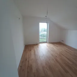 Poddasze z nowo ułożonymi panelami podłogowymi o ciepłym, dębowym odcieniu, świeżo pomalowane na biało ściany, widoczne niedokończone instalacje elektryczne i okno balkonowe z widokiem na zieleń.