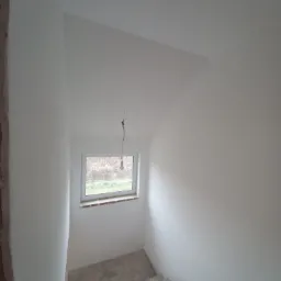 Wnętrze klatki schodowej w trakcie remontu, ściany pomalowane na biało, widoczne schody w stanie surowym, okno z widokiem na zewnątrz i wisząca żarówka na kablu.