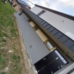 Nowoczesna elewacja domu jednorodzinnego w odcieniach szarości i bieli, z elementami drewnopodobnymi i czarnymi detalami, widok z dołu, częściowo wykończony teren wokół budynku.