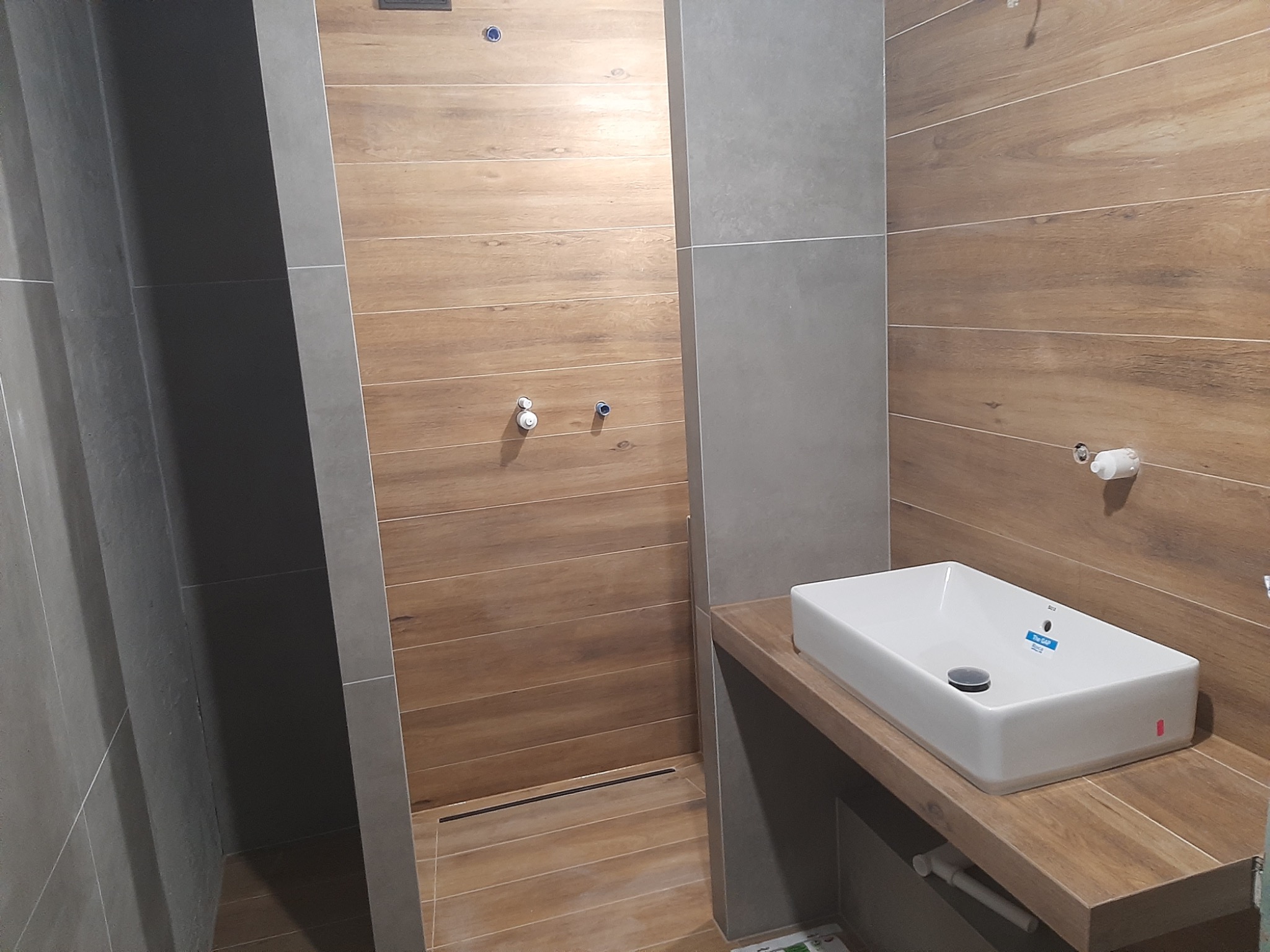 Nowoczesna łazienka z drewnopodobnymi i szarymi płytkami, białą prostokątną umywalką nablatową marki Roca, odpływem liniowym w kabinie prysznicowej bez brodzika i wystającymi rurami instalacji wodnej.