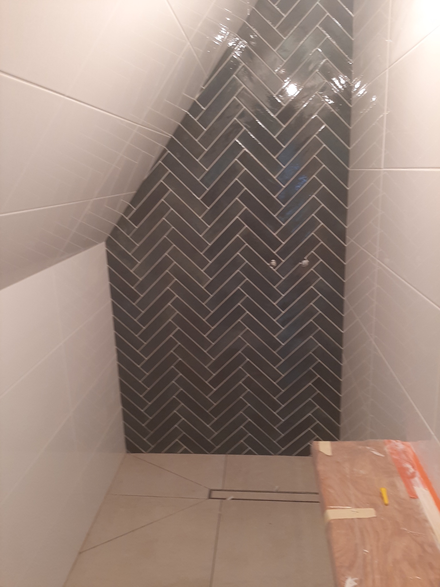 Wykończona łazienka na poddaszu z szarymi płytkami ułożonymi w jodełkę na ścianie prysznicowej i białymi płytkami na pozostałych ścianach, widoczny odpływ liniowy i zabezpieczona folią ochronną wanna.