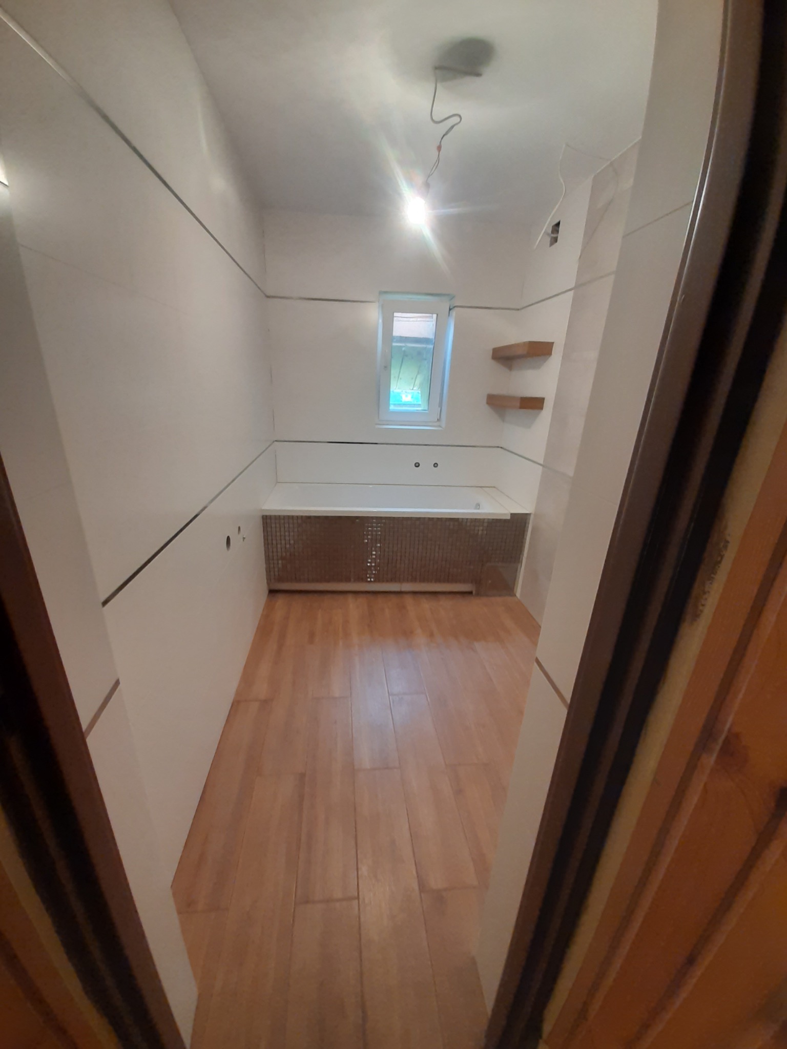 Wnętrze łazienki w trakcie wykańczania, widok zza uchylonych drewnianych drzwi, z widoczną wanną obudowaną mozaiką, drewnianą podłogą i dwoma półkami na ścianie.