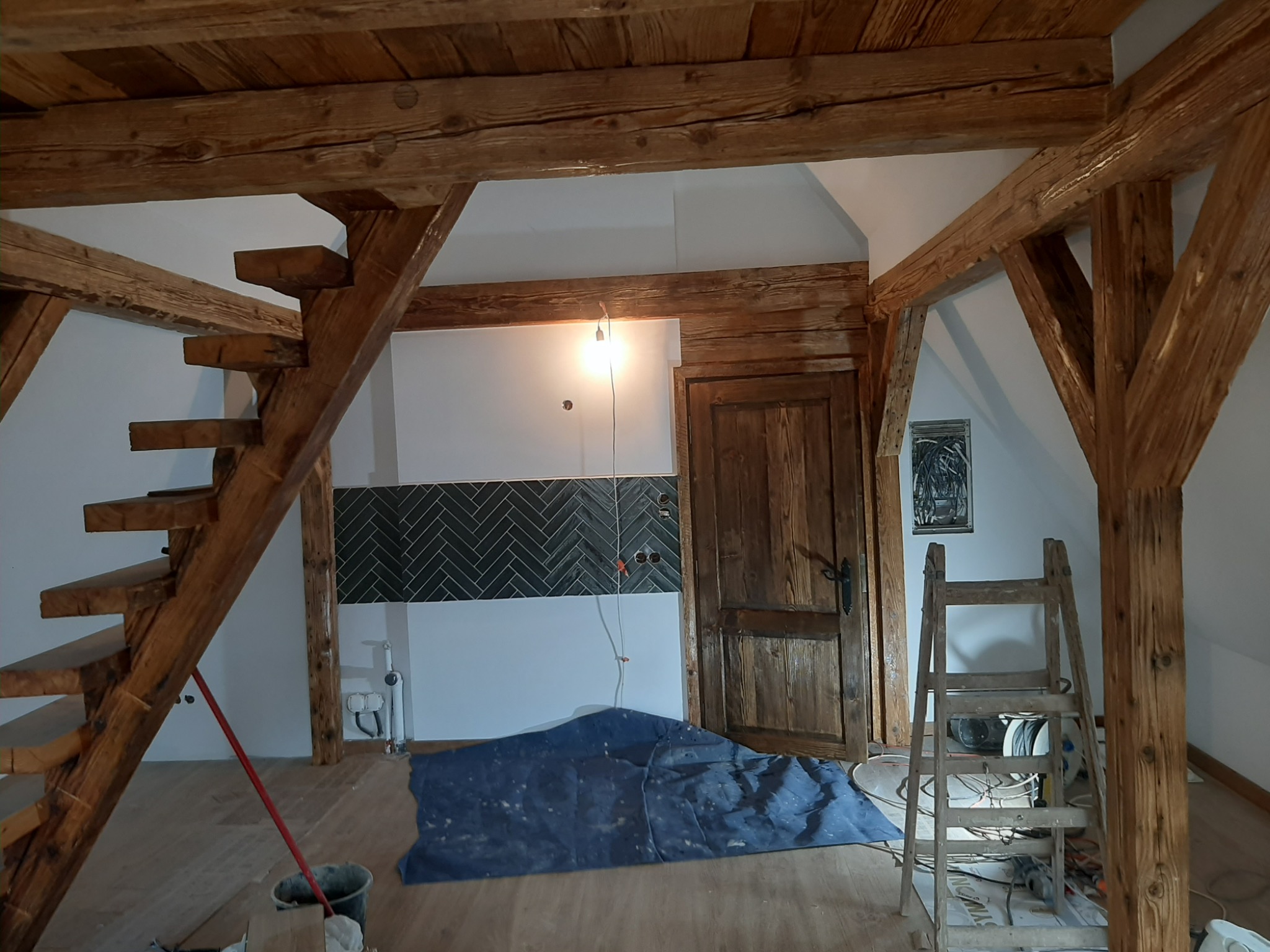 Poddasze w trakcie remontu: drewniane belki konstrukcyjne, schody i drzwi, fragment ściany z ciemną glazurą ułożoną w jodełkę, zabezpieczona podłoga, drabina malarska i widoczny bałagan budowlany.